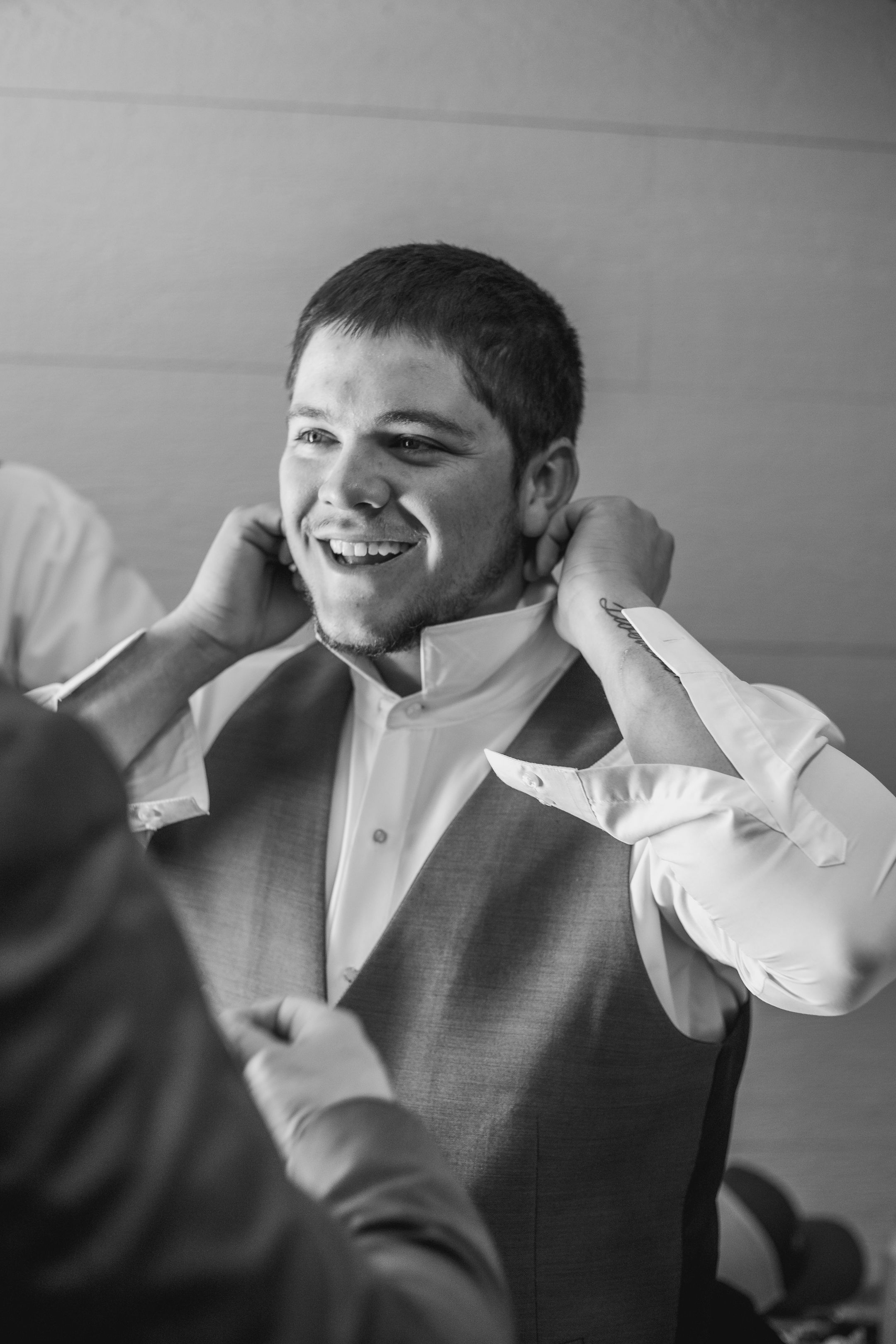 A person in a white dress shirt and gray vest smiles while being helped with formal attire preparations.