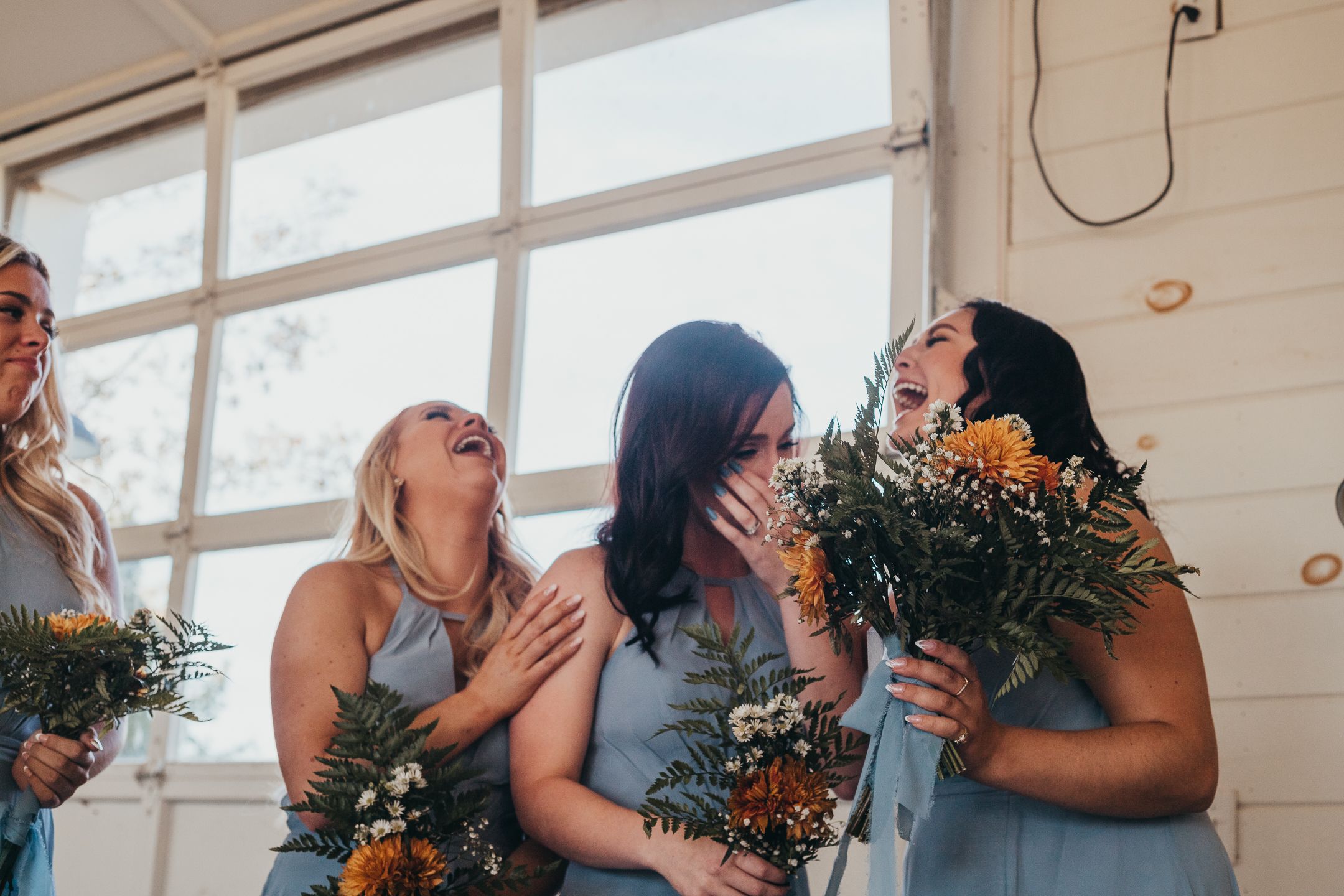 A group of bridesmaids in light blue dresses laugh together while holding orange and green bouquets.