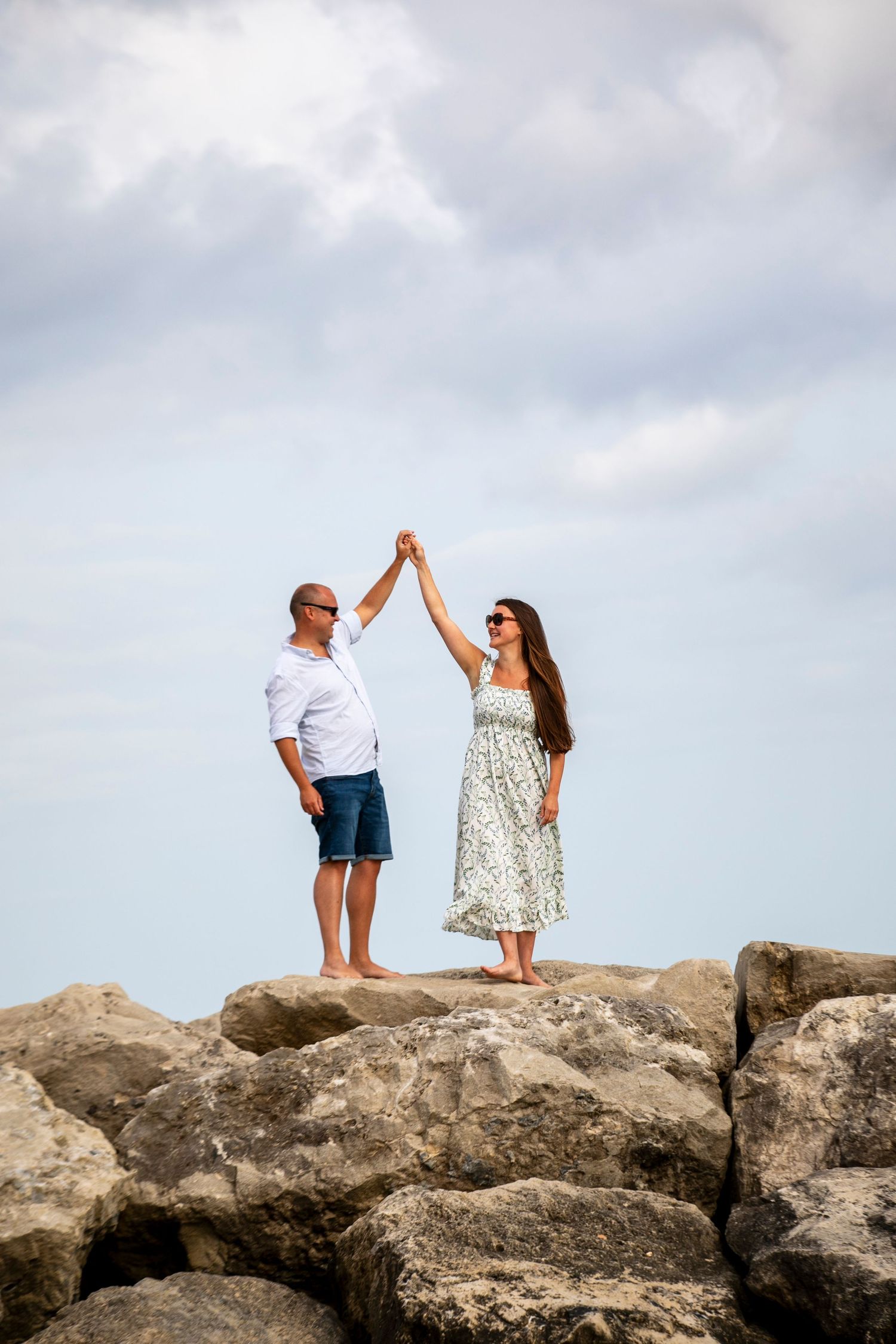 A couple poses playfully on large coastal rocks with cloudy skies in the background.