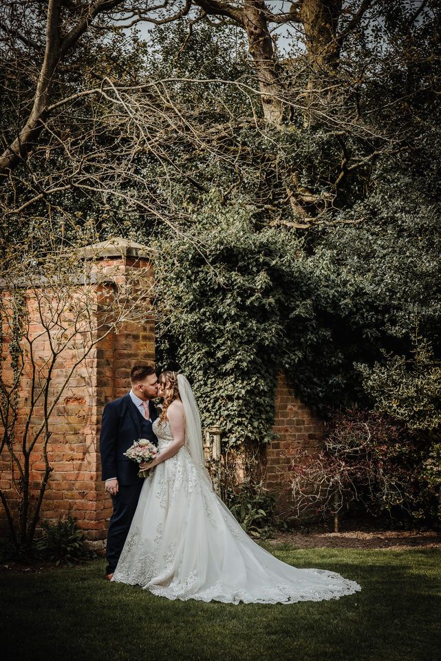 A wedding couple stands together by an old brick wall in a garden setting with moody lighting and trailing vines.