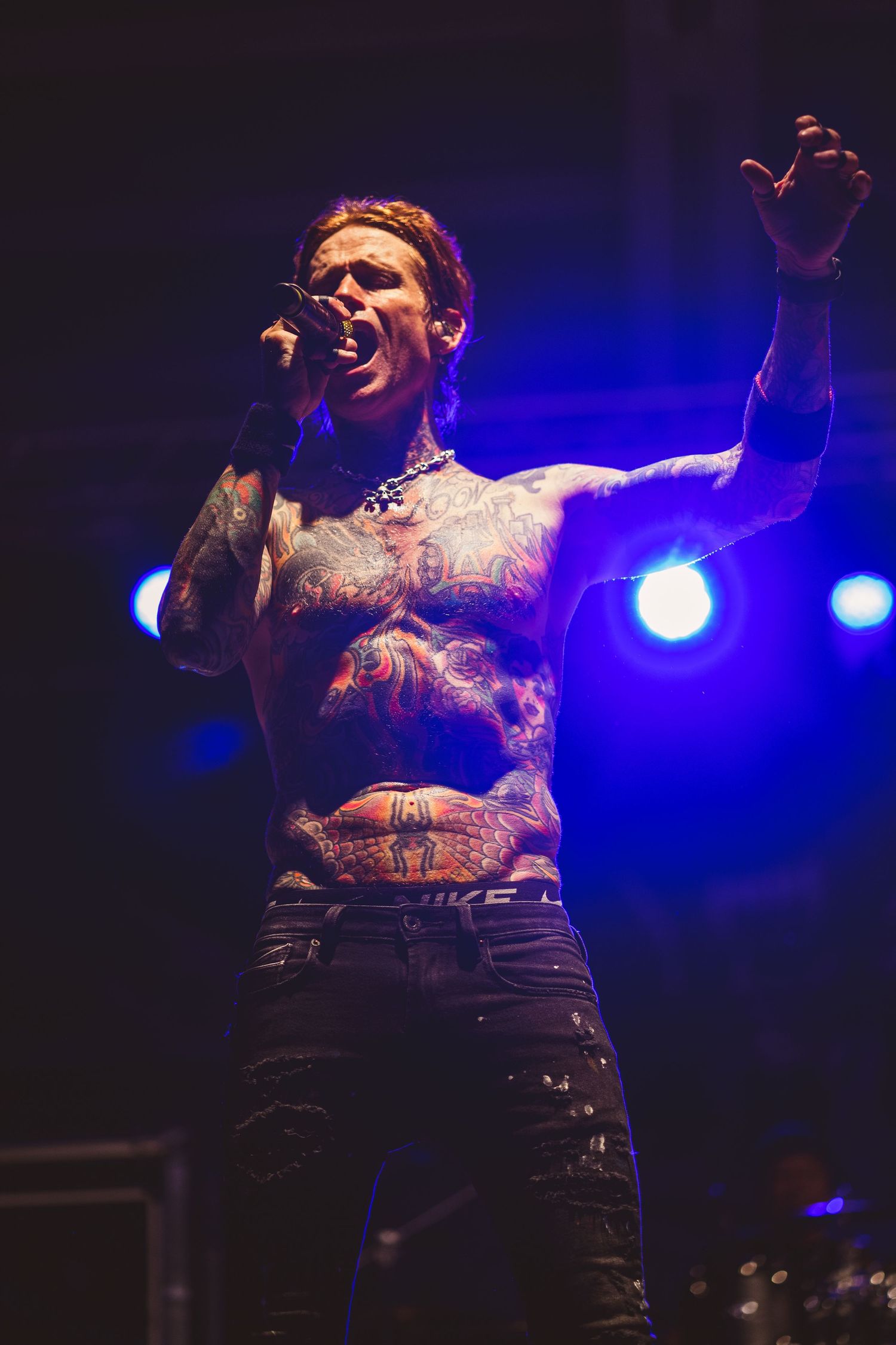 Performer with tattoos singing emotionally on stage with blue concert lighting in the background.