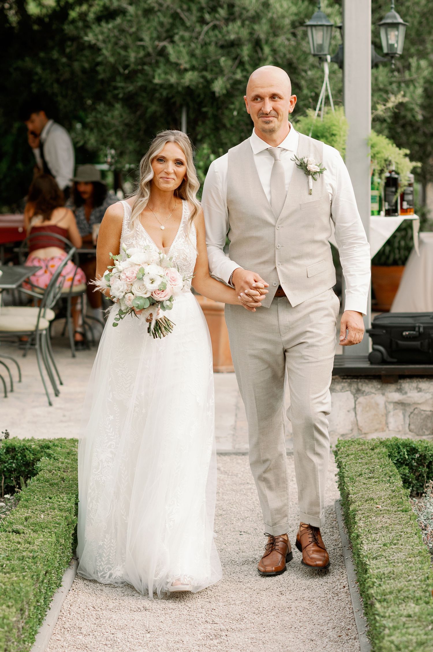 A bride and groom walking together on a garden path in light-colored wedding attire.