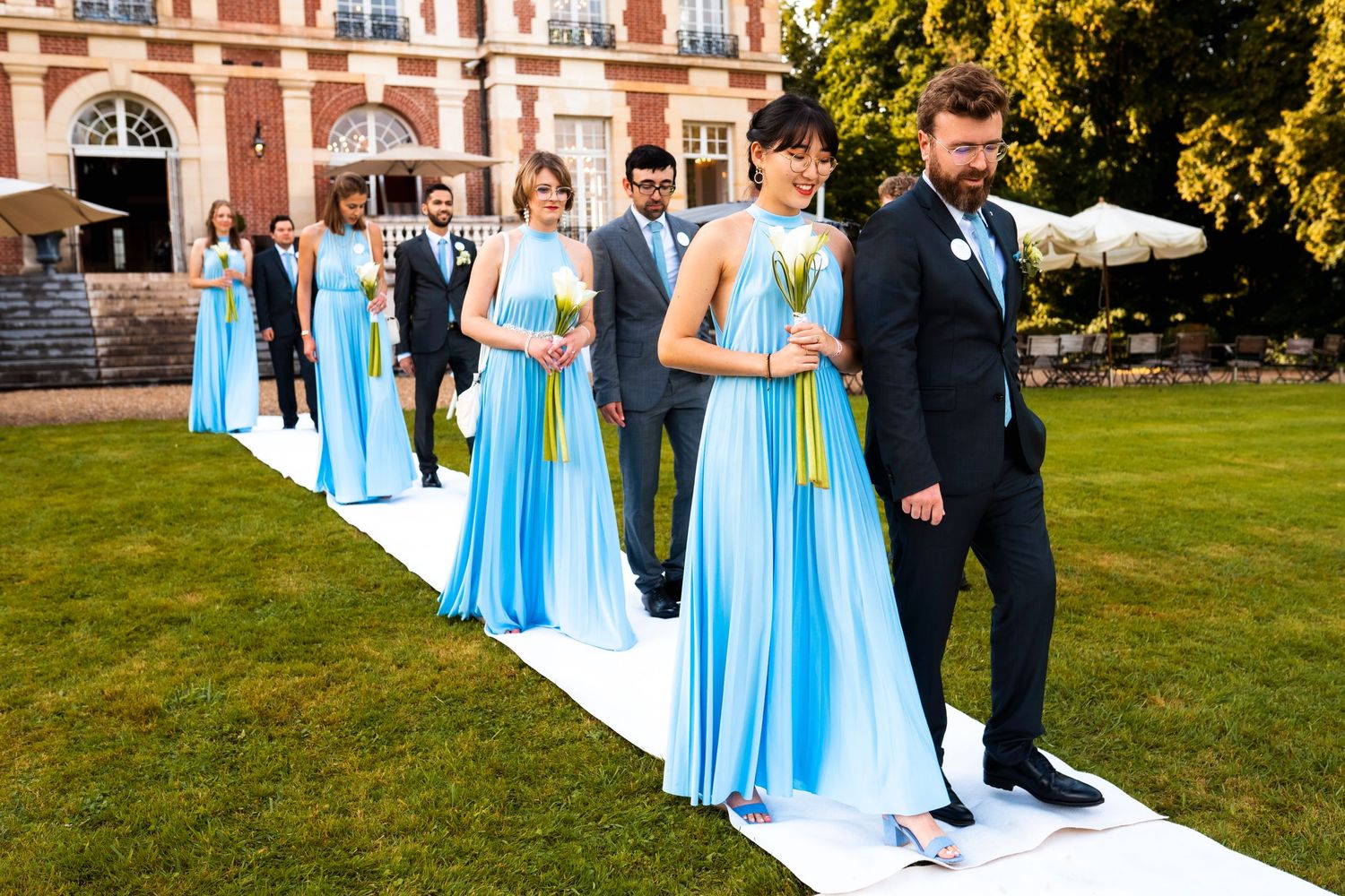 Wedding party members in light blue dresses and dark suits walking on white carpet outdoors.