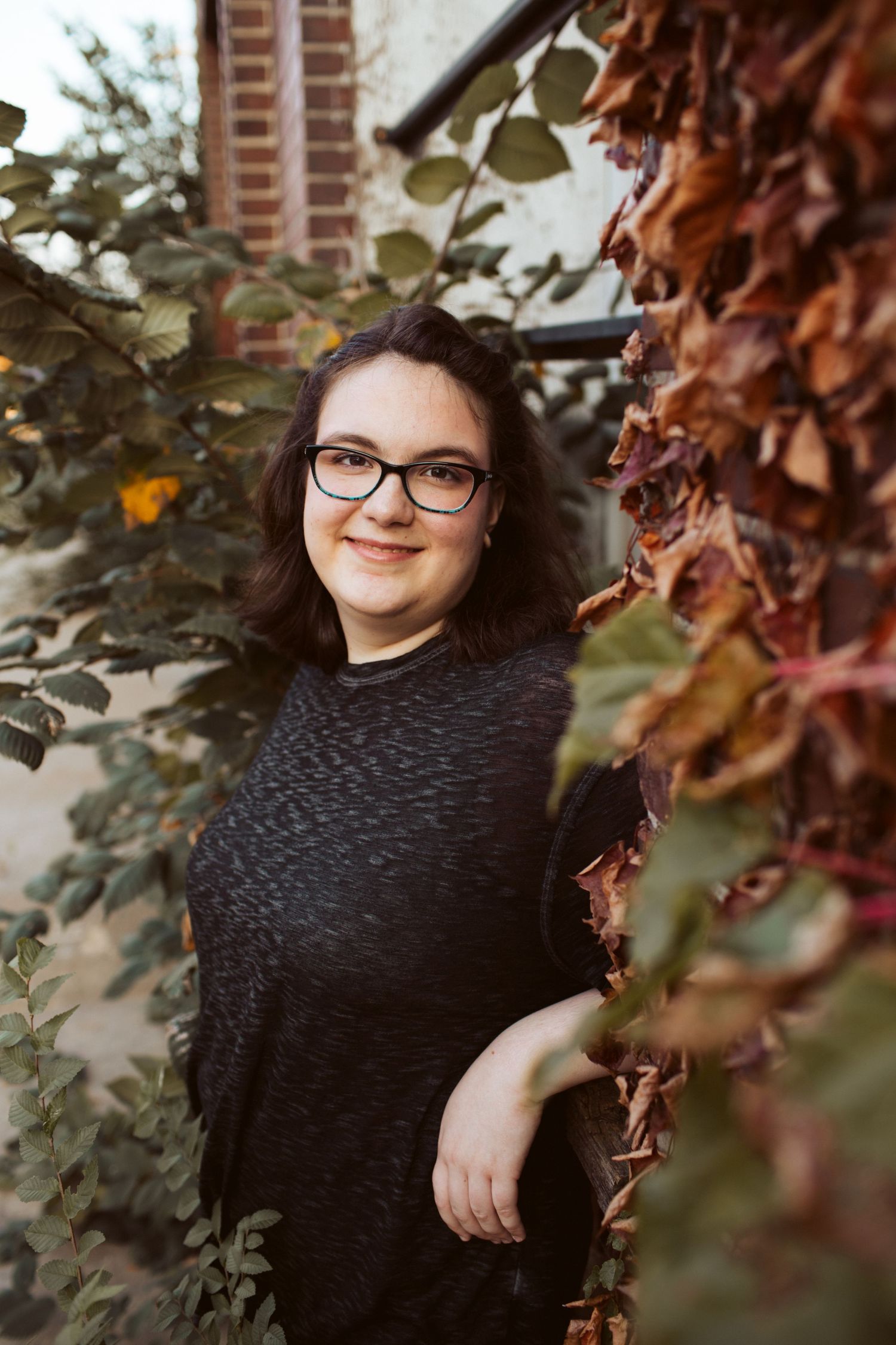 Smiling young woman with glasses in dark sweater poses among autumn foliage near brick building exterior.