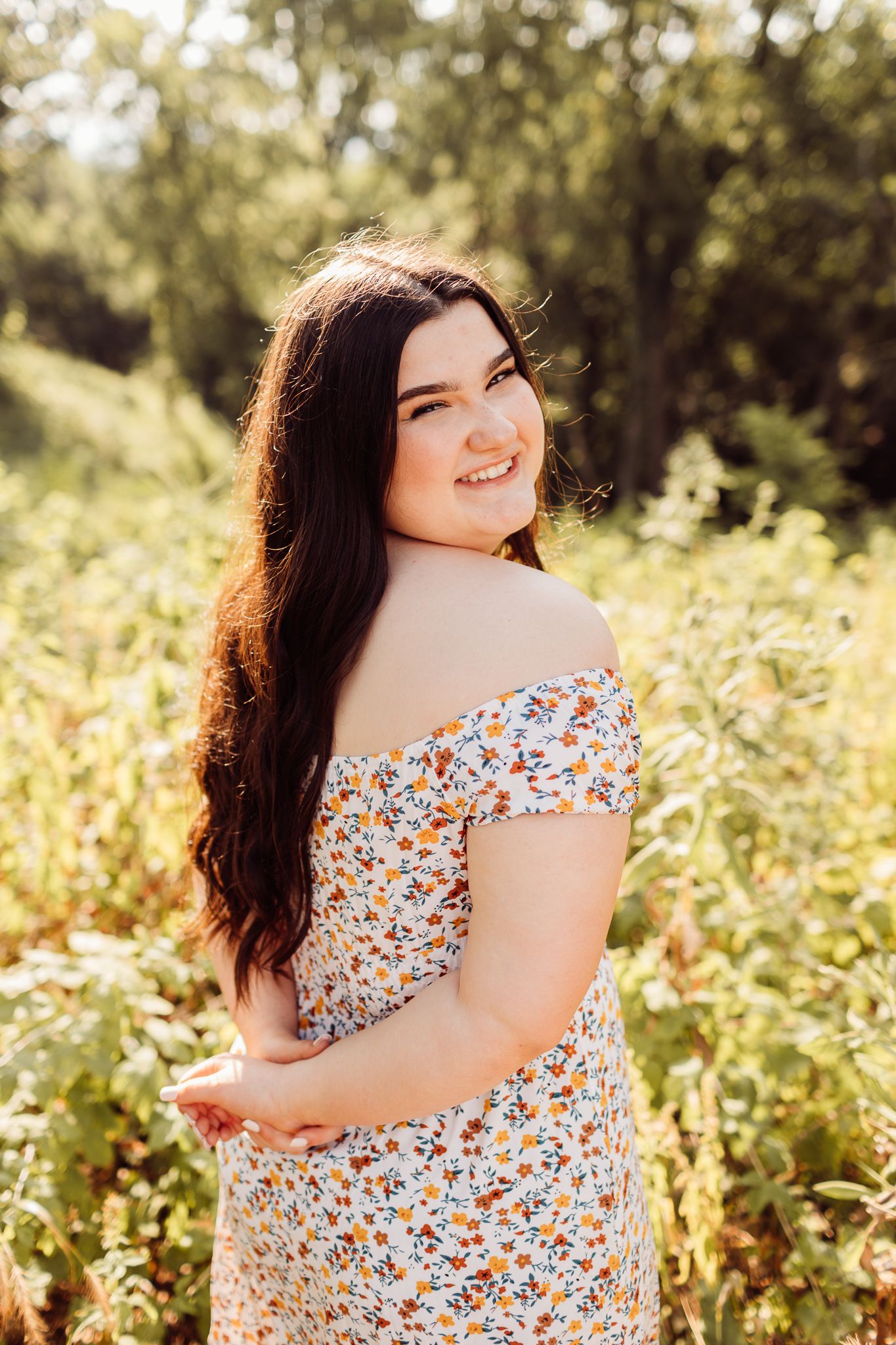 Smiling young woman in a floral off-shoulder dress posing in a sunlit green outdoor setting.