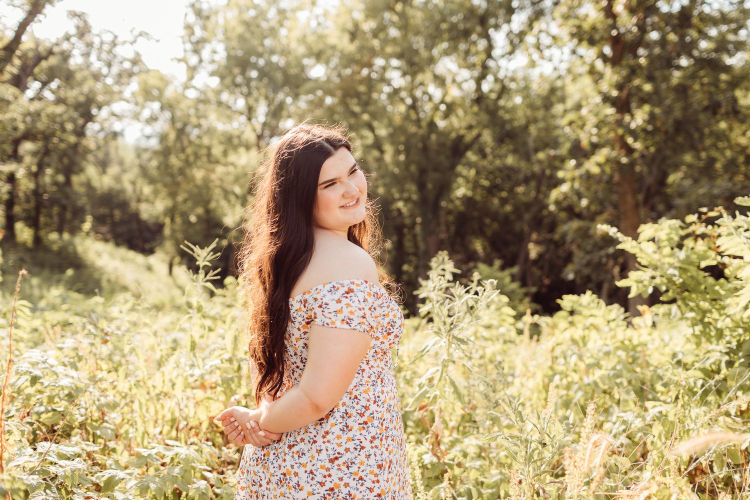 Young woman with long dark hair in a floral off-shoulder dress standing in a sunny wildflower field.