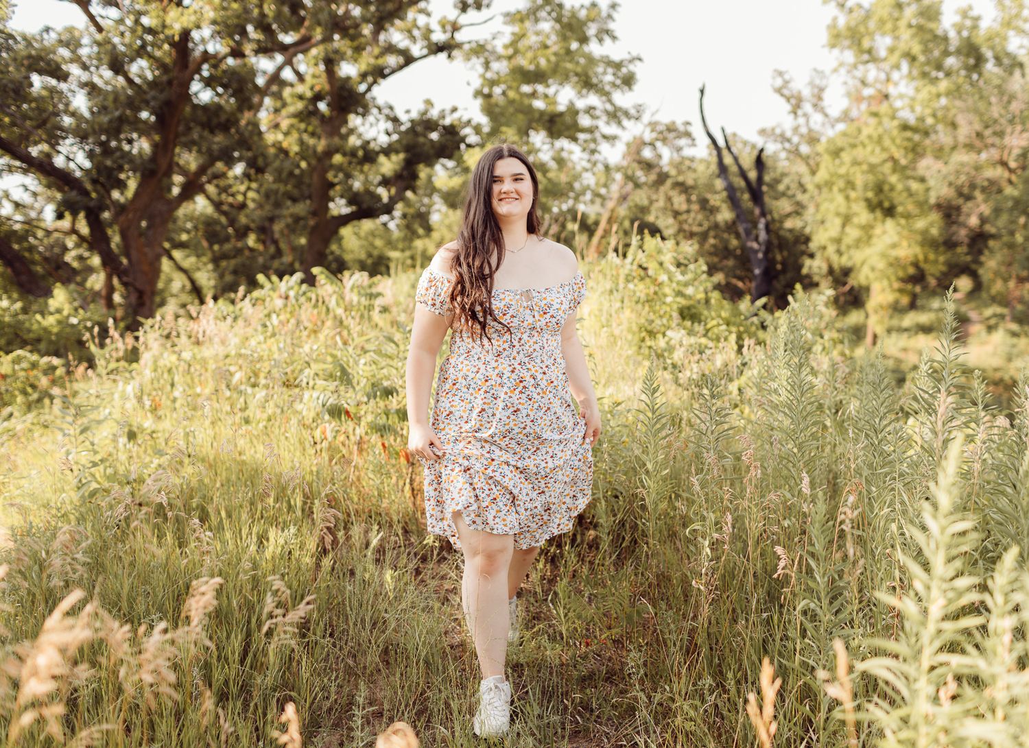 Young woman in a floral sundress walking through tall golden grass in a sunny natural field.