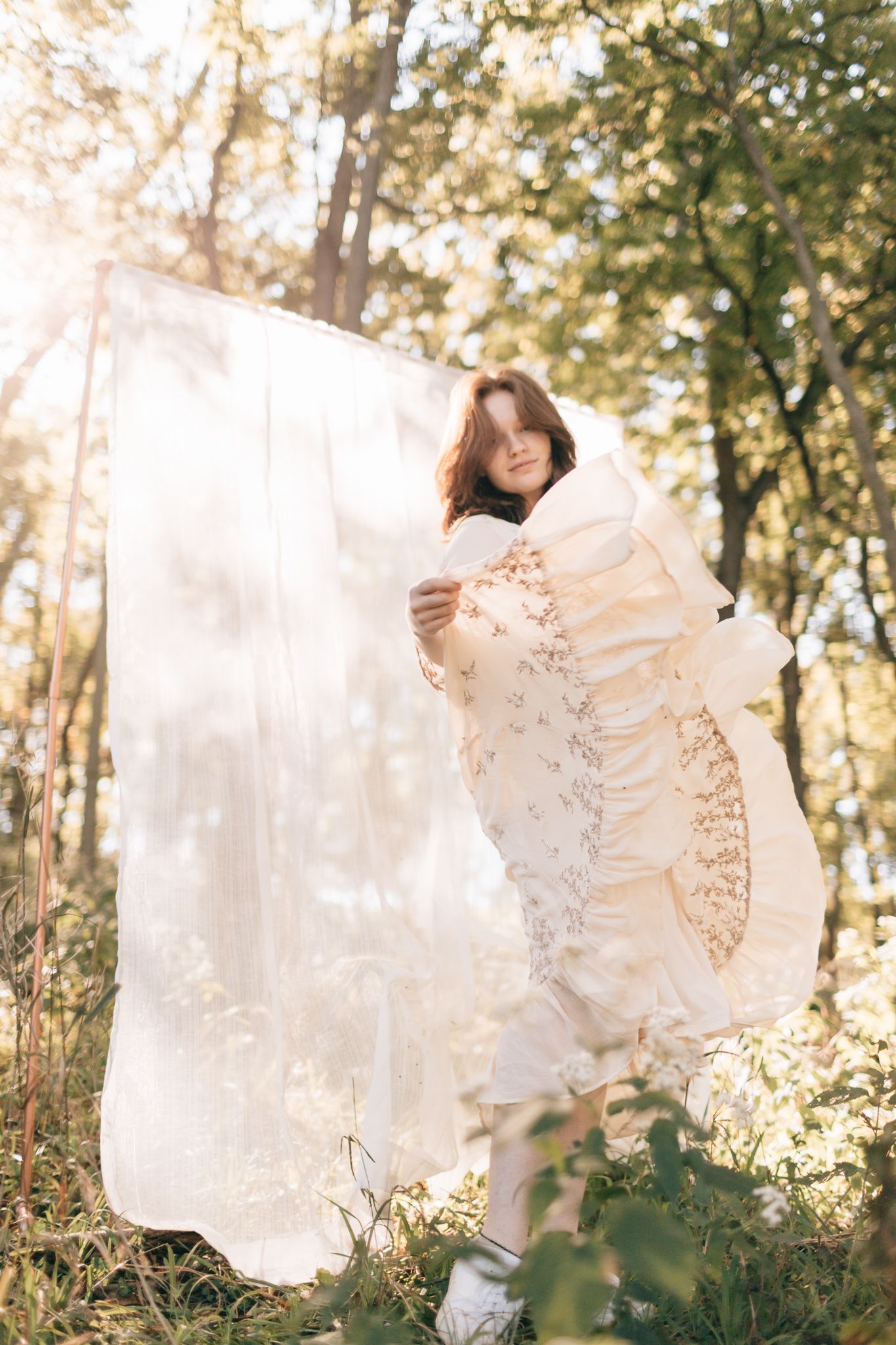 Woman in elegant ivory lace dress wraps herself in sheer white fabric among sunlit trees in a forest.