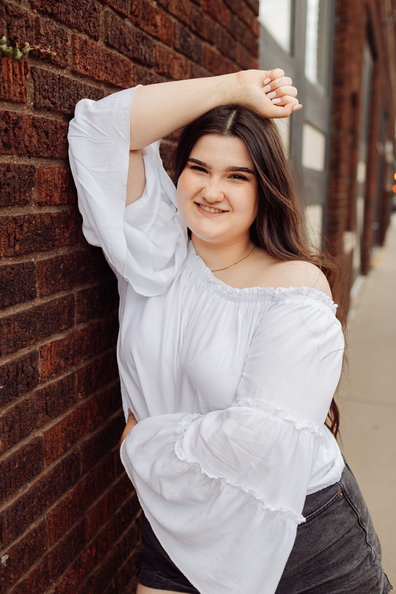 Smiling young woman in a white off-shoulder top leaning against a red brick wall outdoors.