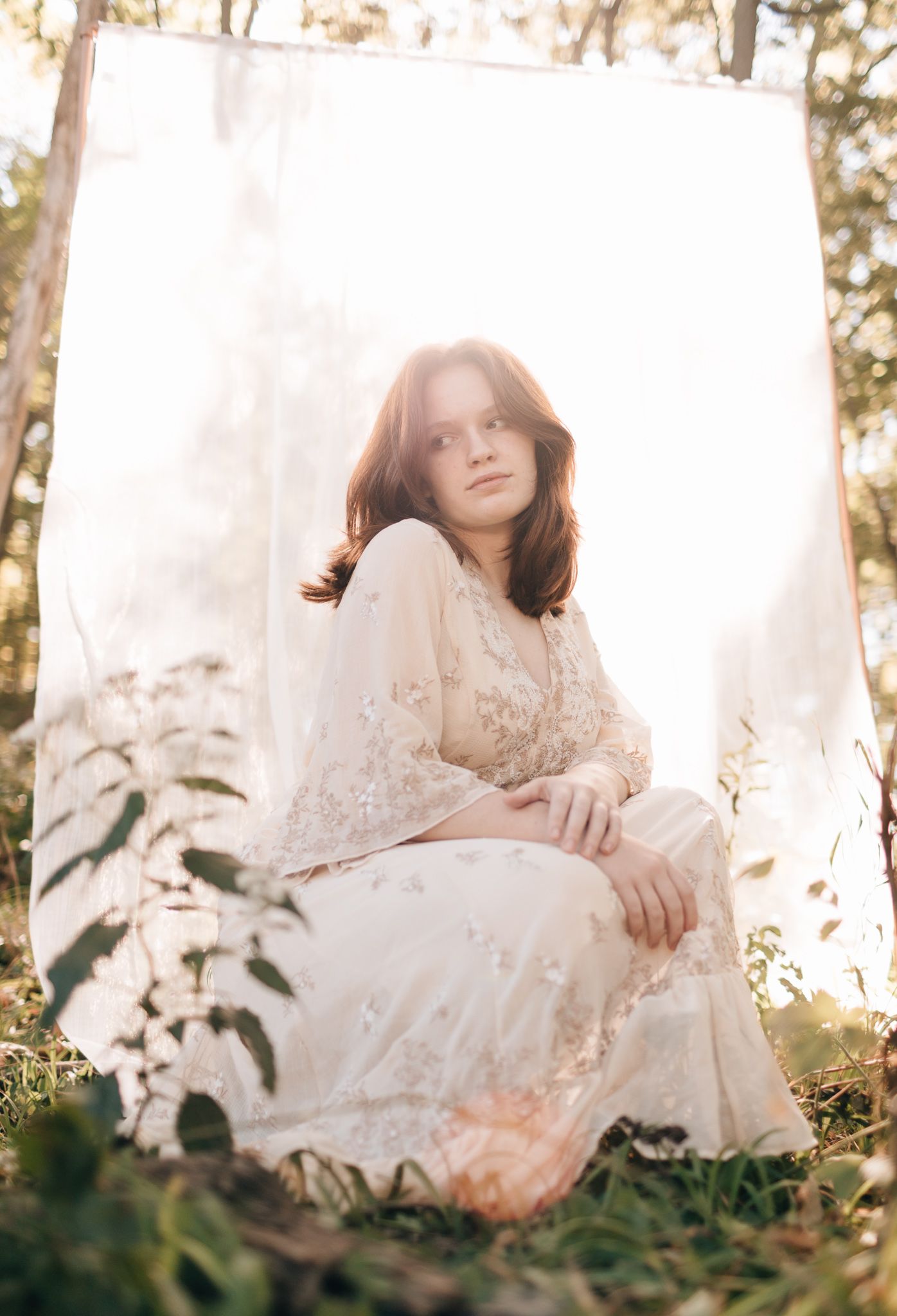 Young woman in floral lace cream dress sits gracefully outdoors against white fabric backdrop in golden sunlight.