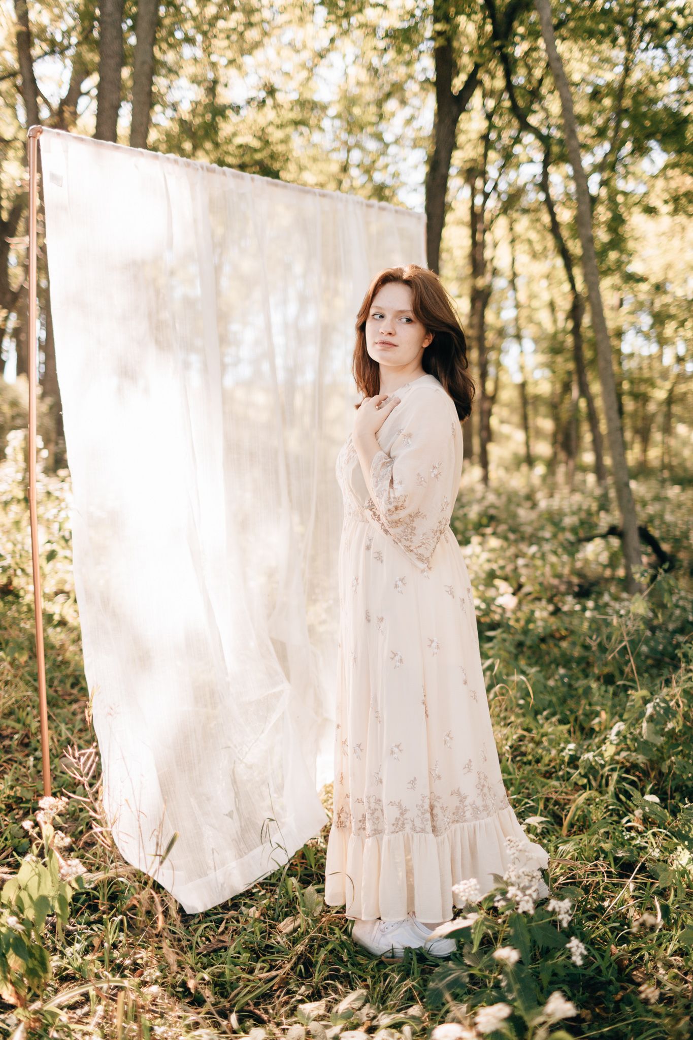 Young woman in vintage cream floral maxi dress stands beside white fabric backdrop in sunny woodland setting.