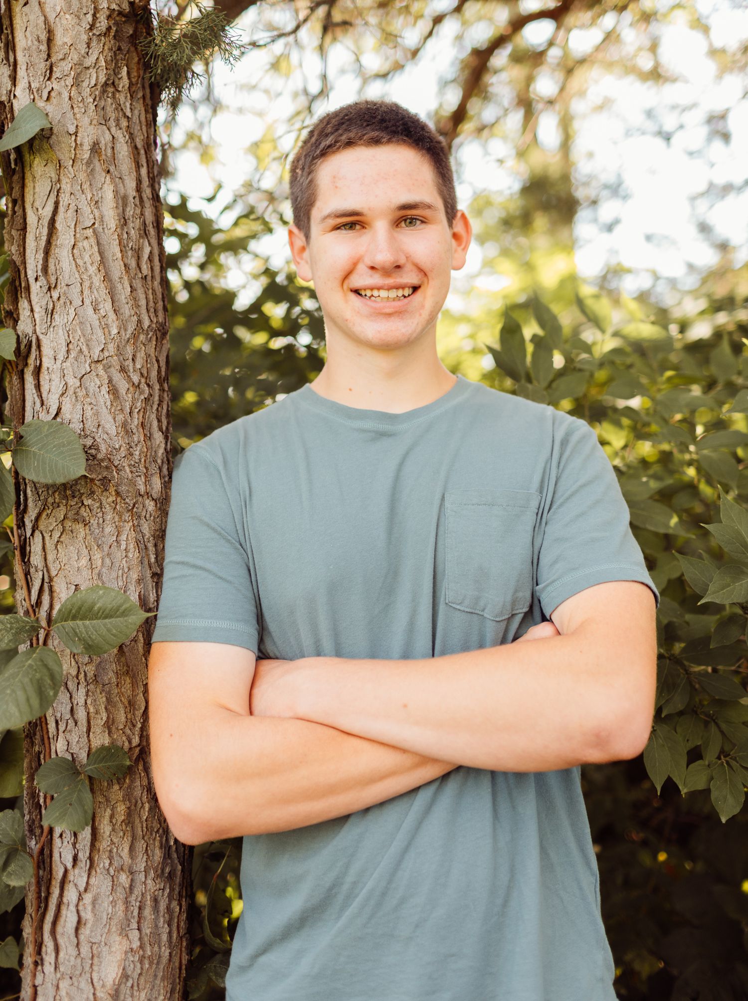 Smiling young man in a gray t-shirt standing with arms crossed beside a tree outdoors.