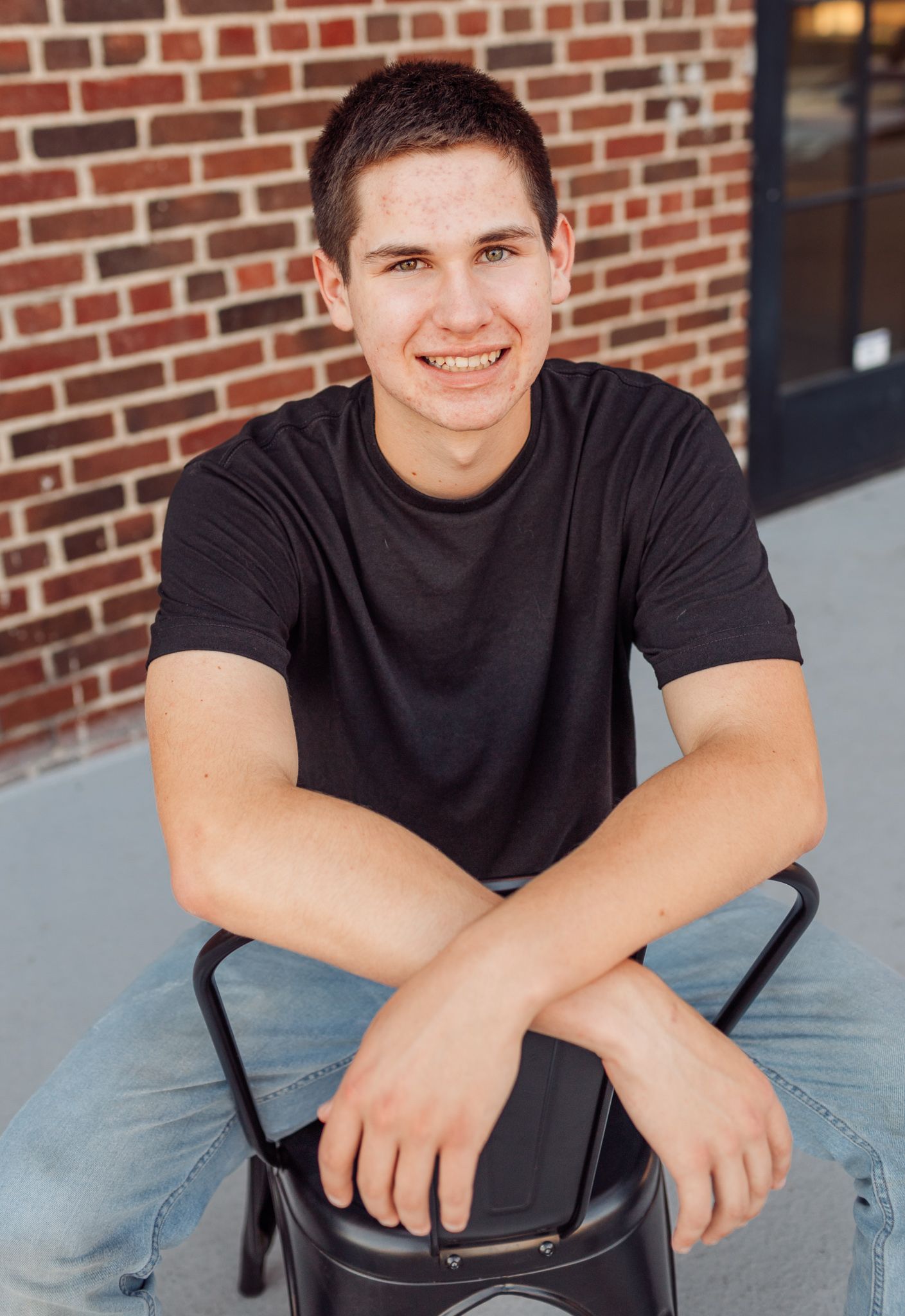 Smiling teenage boy in a black t-shirt sitting on a stool in front of a brick wall.