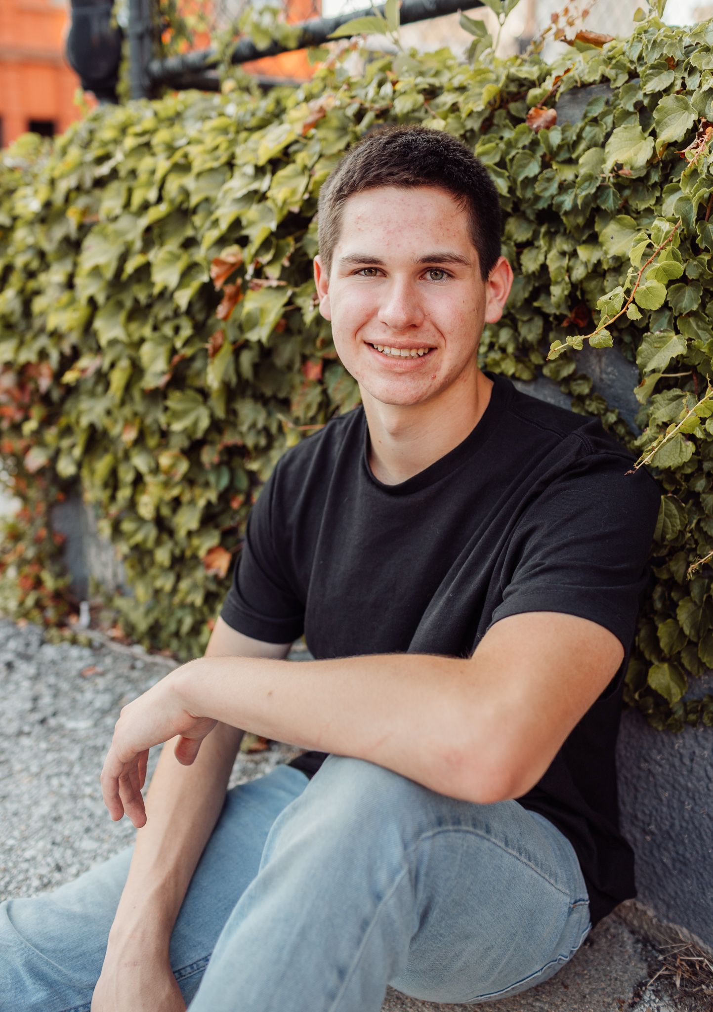 Young man in black t-shirt and jeans sitting outdoors against ivy-covered wall, smiling for senior portrait photo.