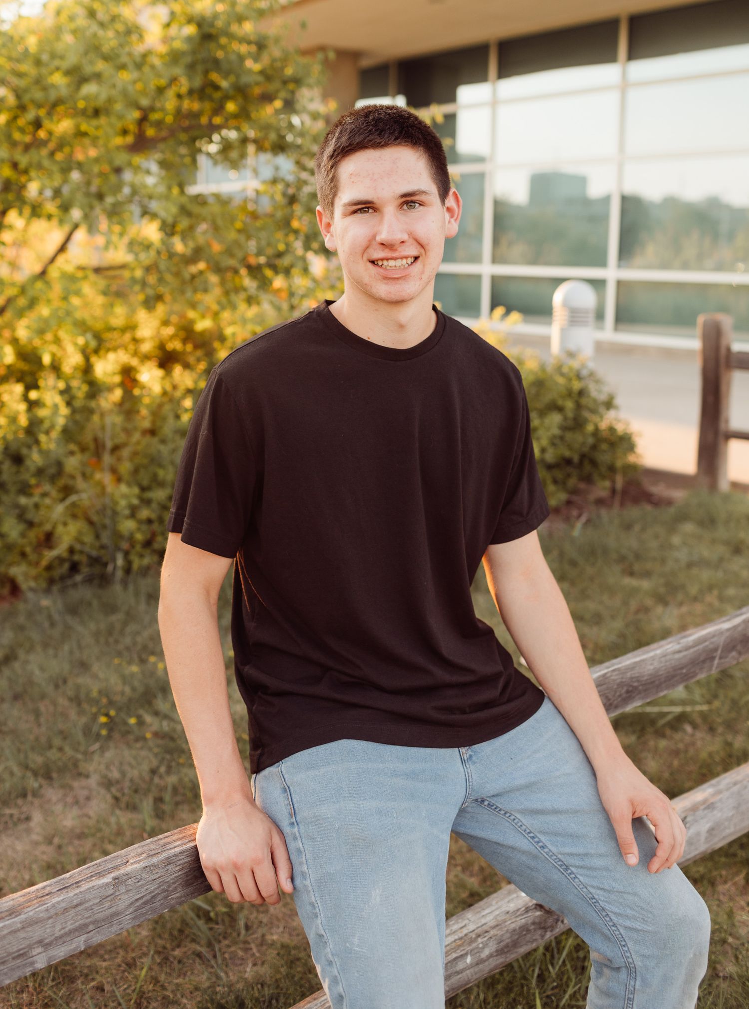 Smiling young man in dark t-shirt and light jeans leaning on wooden fence outdoors for senior portrait.
