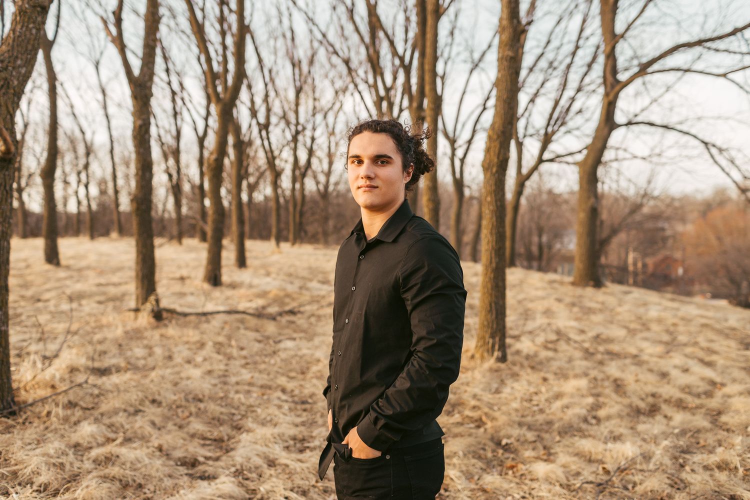 Young man in black shirt standing among bare trees in golden field portrait.