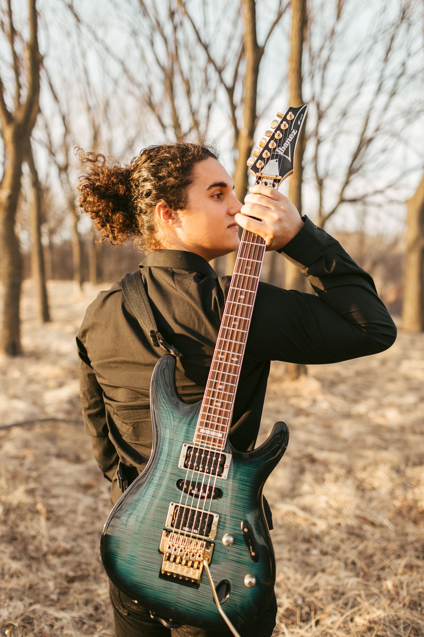 Young musician holding electric guitar upright among bare trees in warm sunset light.