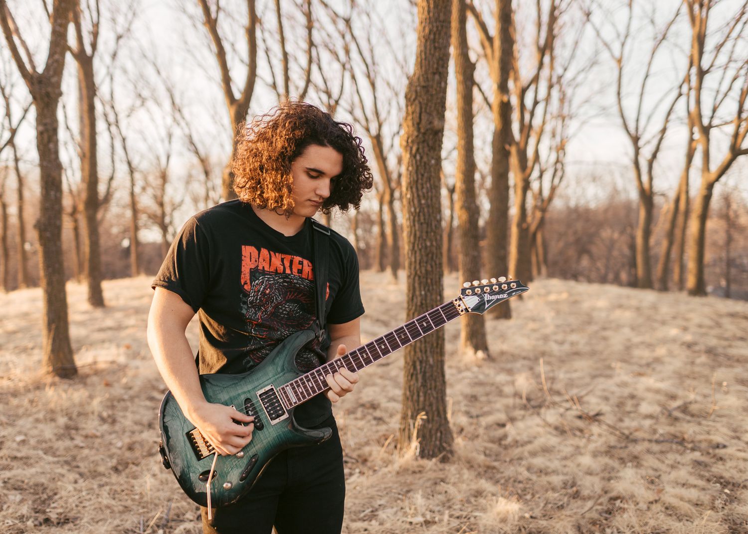 Curly-haired guitarist playing electric guitar outdoors among bare trees in natural light.