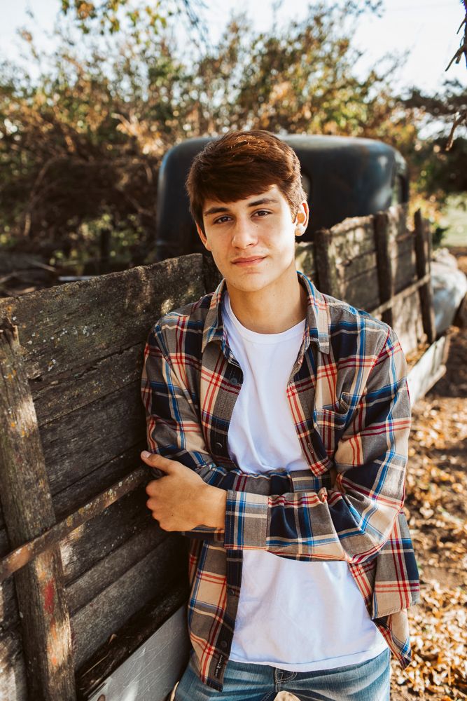 Teen boy in plaid flannel shirt with arms crossed leans against rustic wooden fence in golden sunlight.