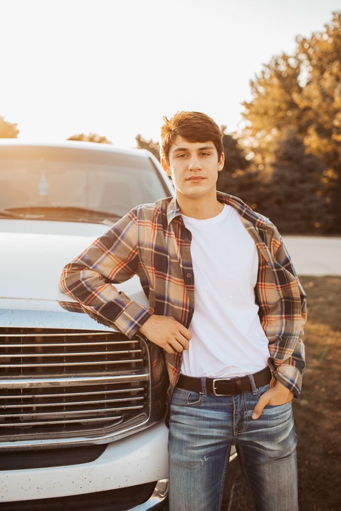 Young man in plaid shirt and jeans leans confidently against white pickup truck in warm sunset light.
