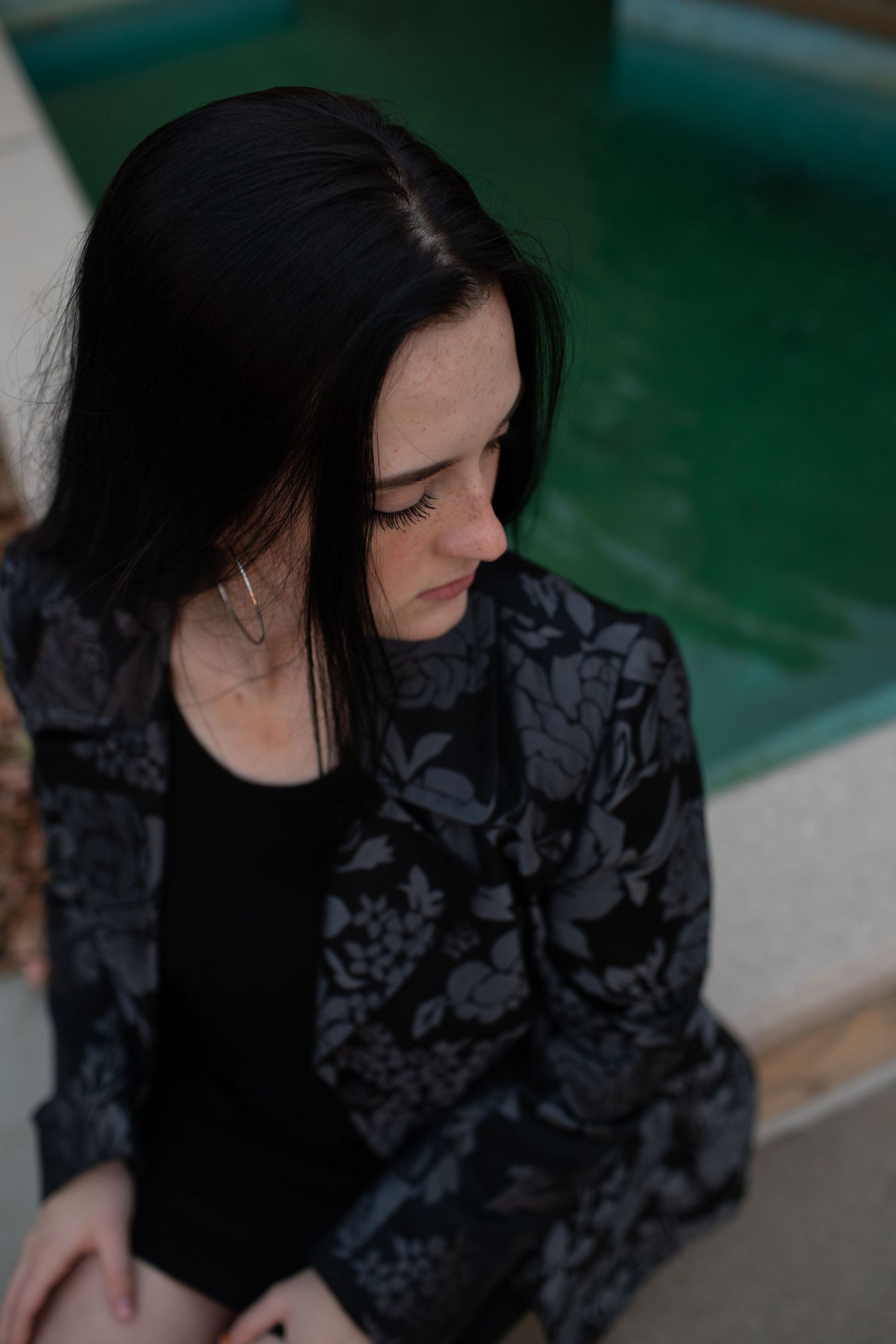 Dark-haired woman in floral blazer sits pensively near turquoise fountain in soft natural lighting.