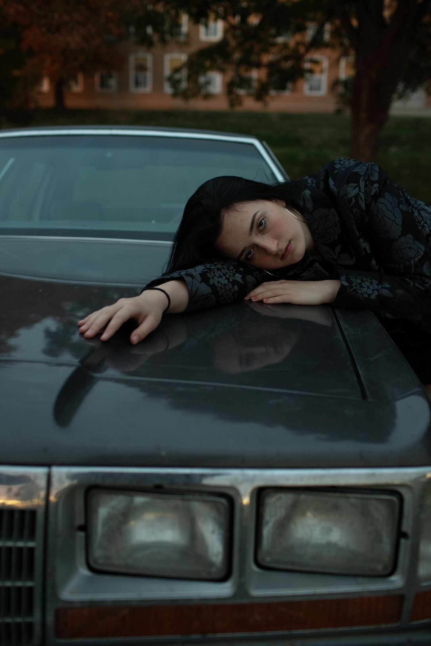 Young dark-haired woman rests her head on the hood of a vintage car in moody evening light.