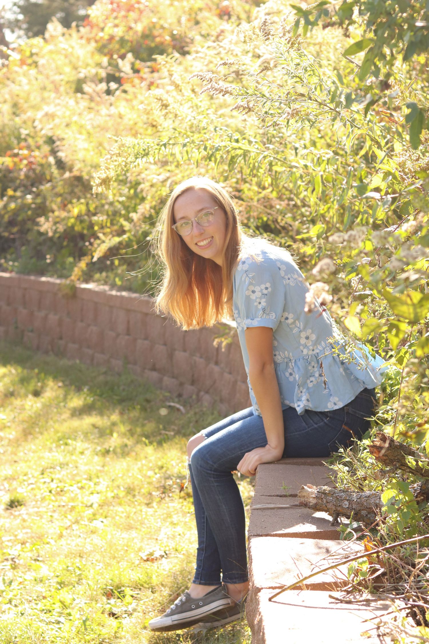 Smiling teenage girl with long red hair sits on garden wall surrounded by lush greenery in sunlight.