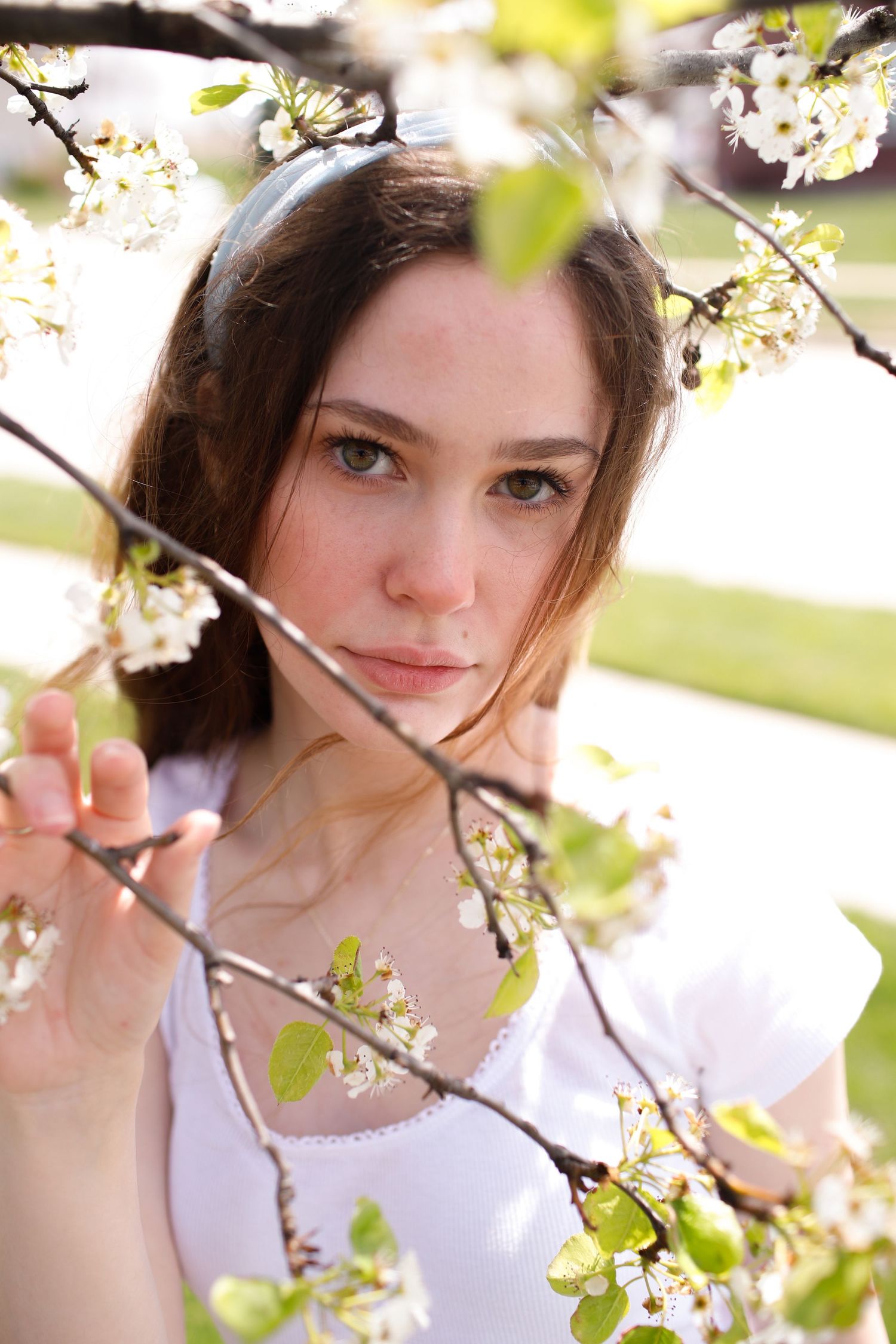 Brunette teen girl gazes through blooming white cherry blossom branches in soft spring daylight.