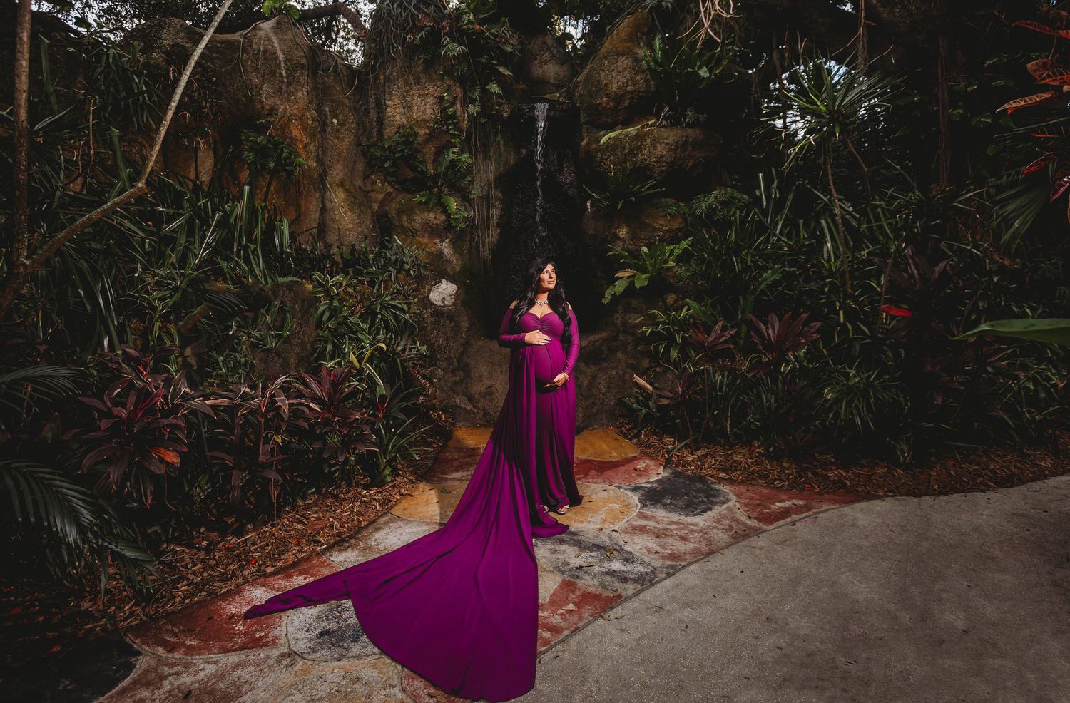 Woman in flowing purple gown poses dramatically on garden path surrounded by lush tropical foliage.
