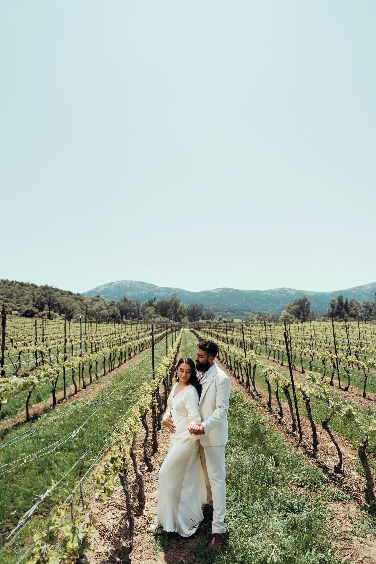 A couple embraces between rows of grapevines in a scenic vineyard.