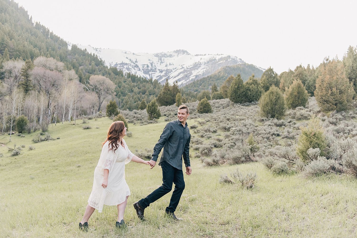 Couple walking hand in hand through mountain meadow with snow-capped peaks in background.