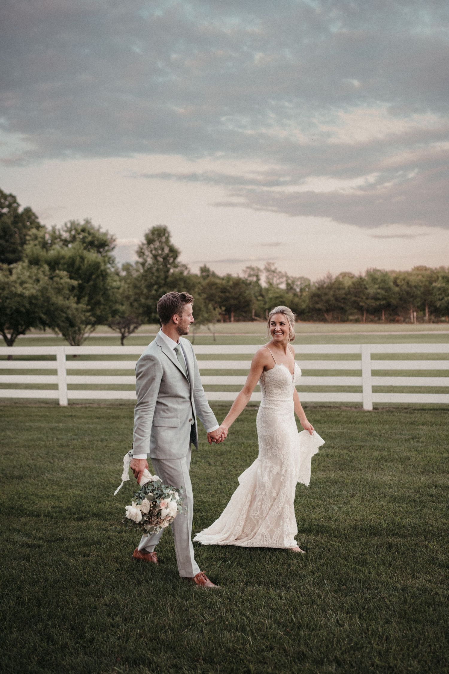 A couple runs playfully across a grassy field with white fences during their wedding celebration.