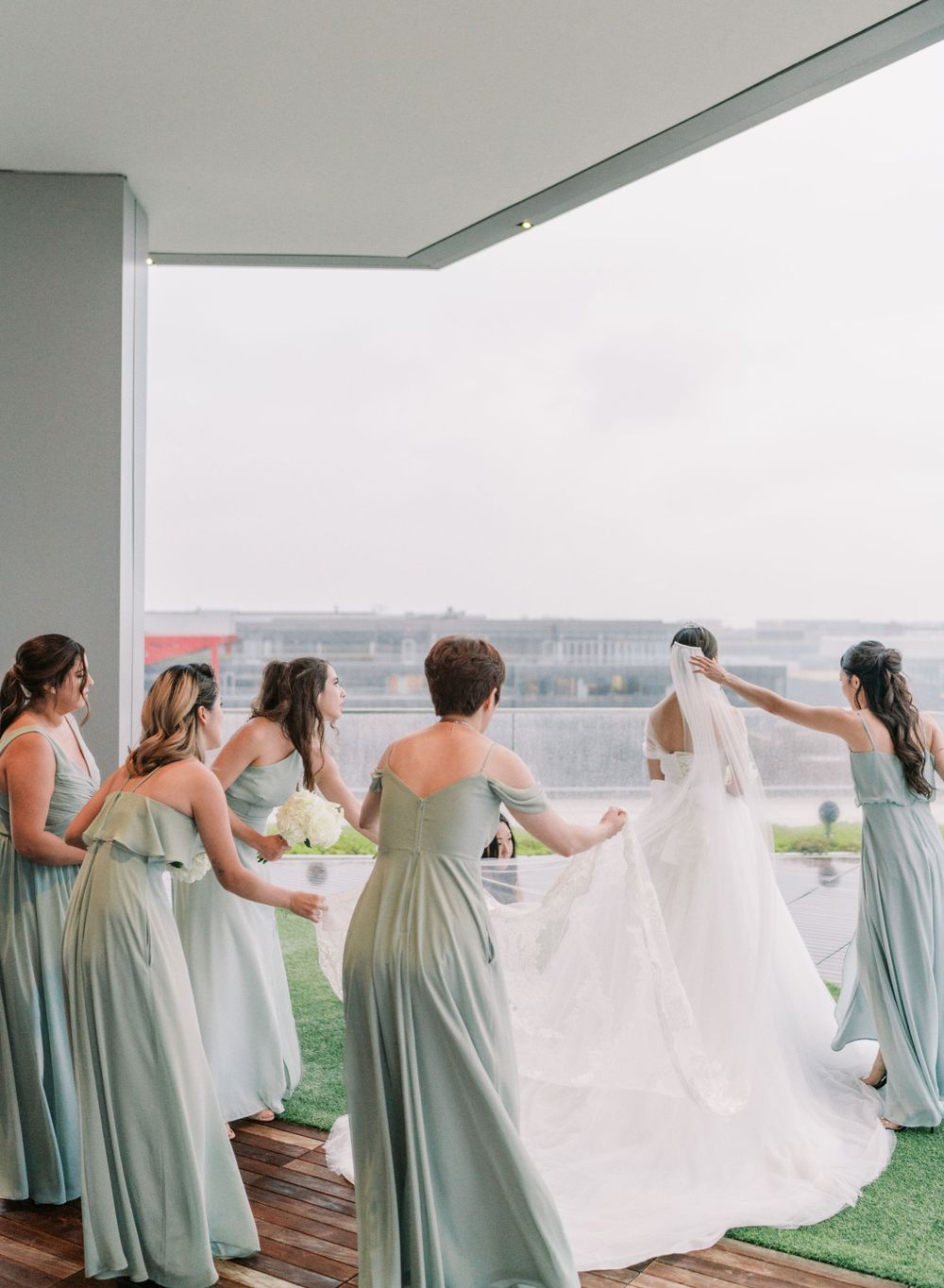 Bridesmaids in sage green dresses help adjust a flowing white wedding gown on an outdoor balcony.