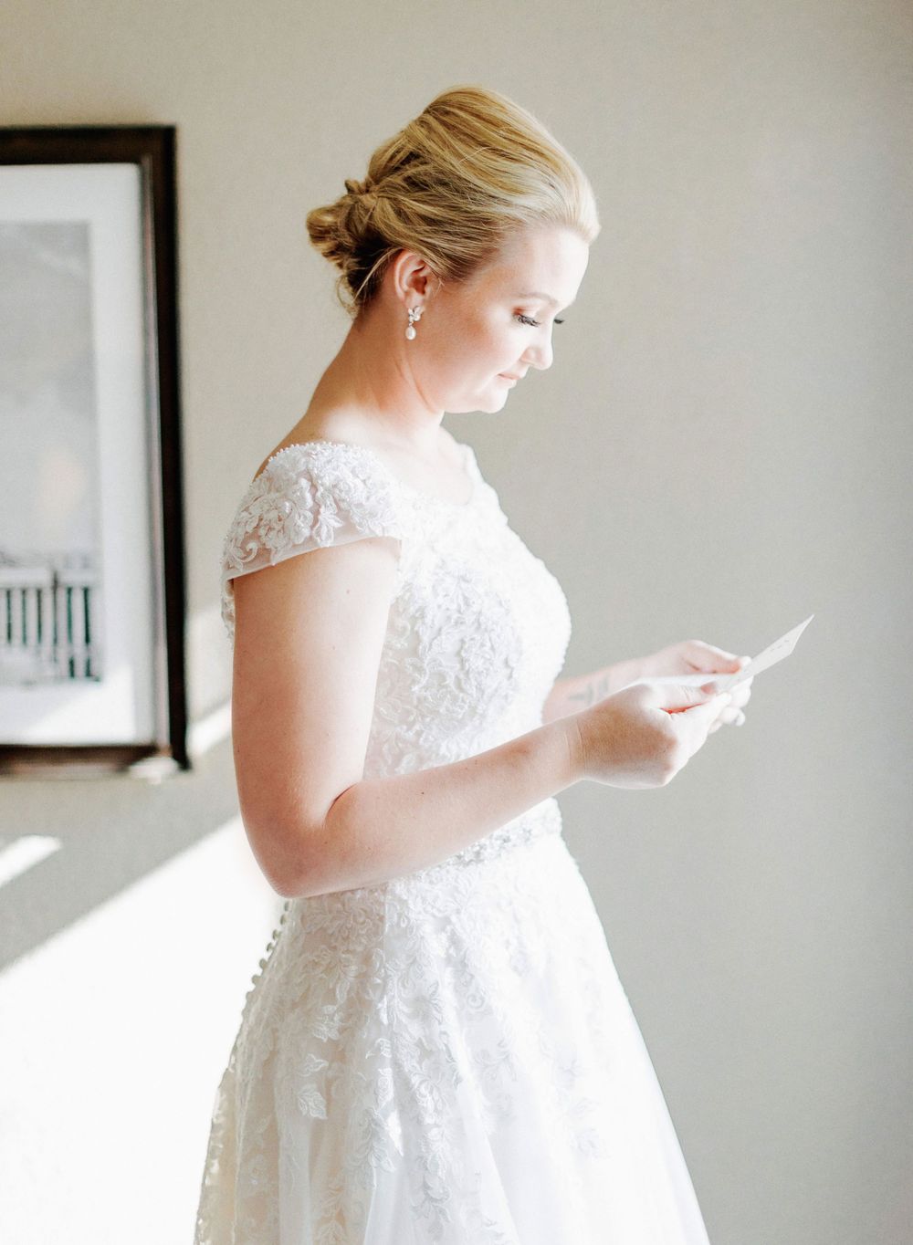 A wedding dress with delicate lace cap sleeves glows in natural window light.