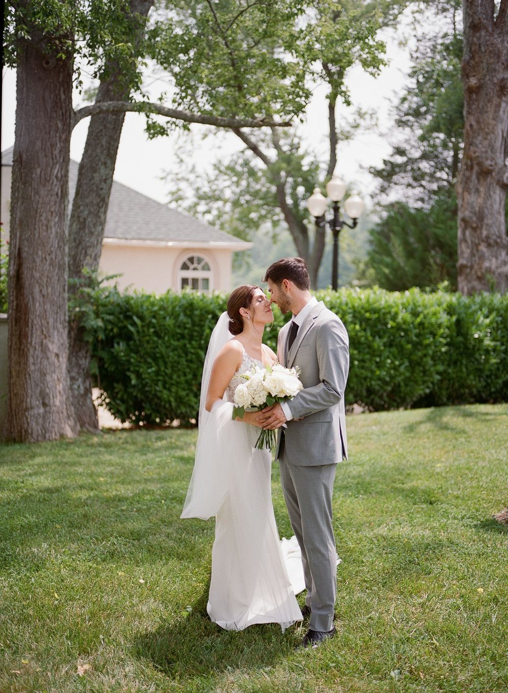 A romantic outdoor wedding portrait with lush greenery and a quaint house in the background.