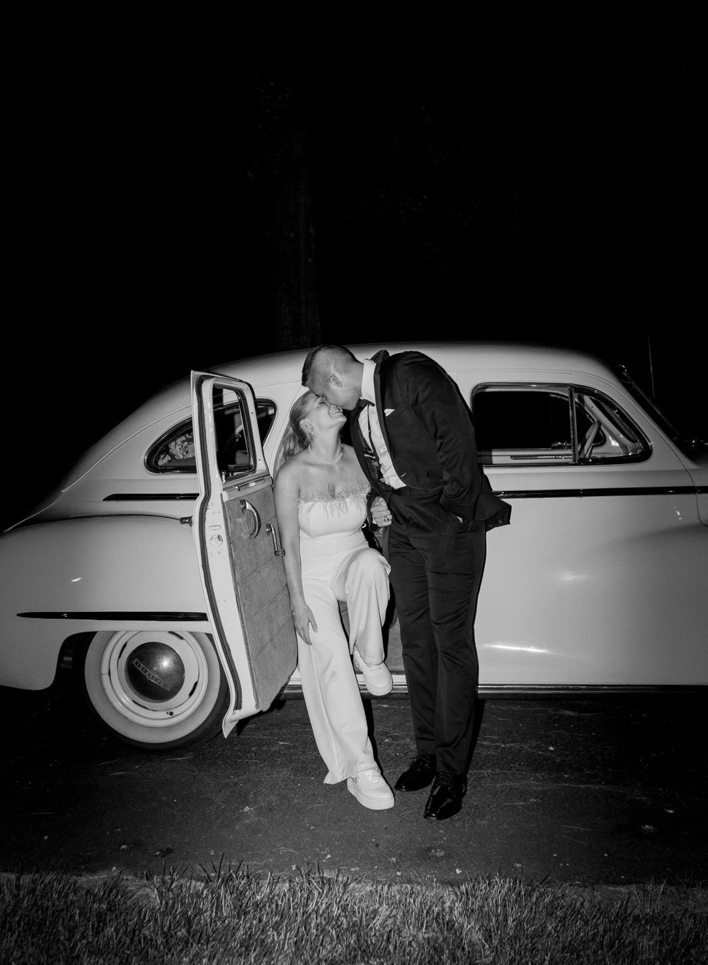 Black and white romantic portrait of newlyweds beside a vintage car at night.