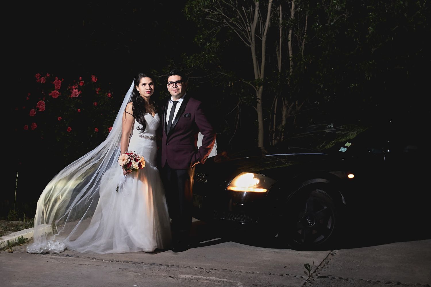 Elegant couple poses beside a vintage car at night after their wedding ceremony.