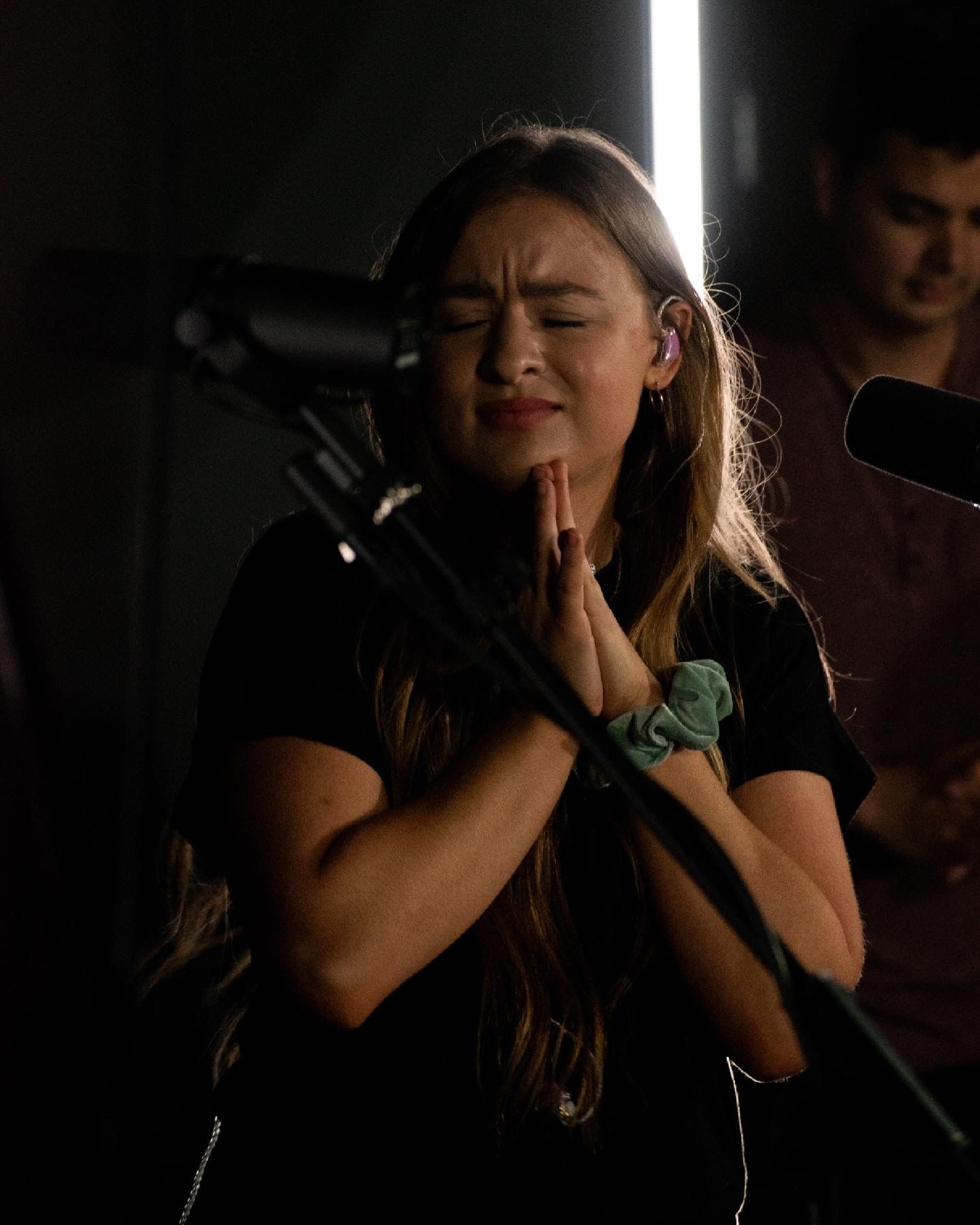 Young woman with hands clasped in prayer, eyes closed, performing on stage with microphone and studio lighting.
