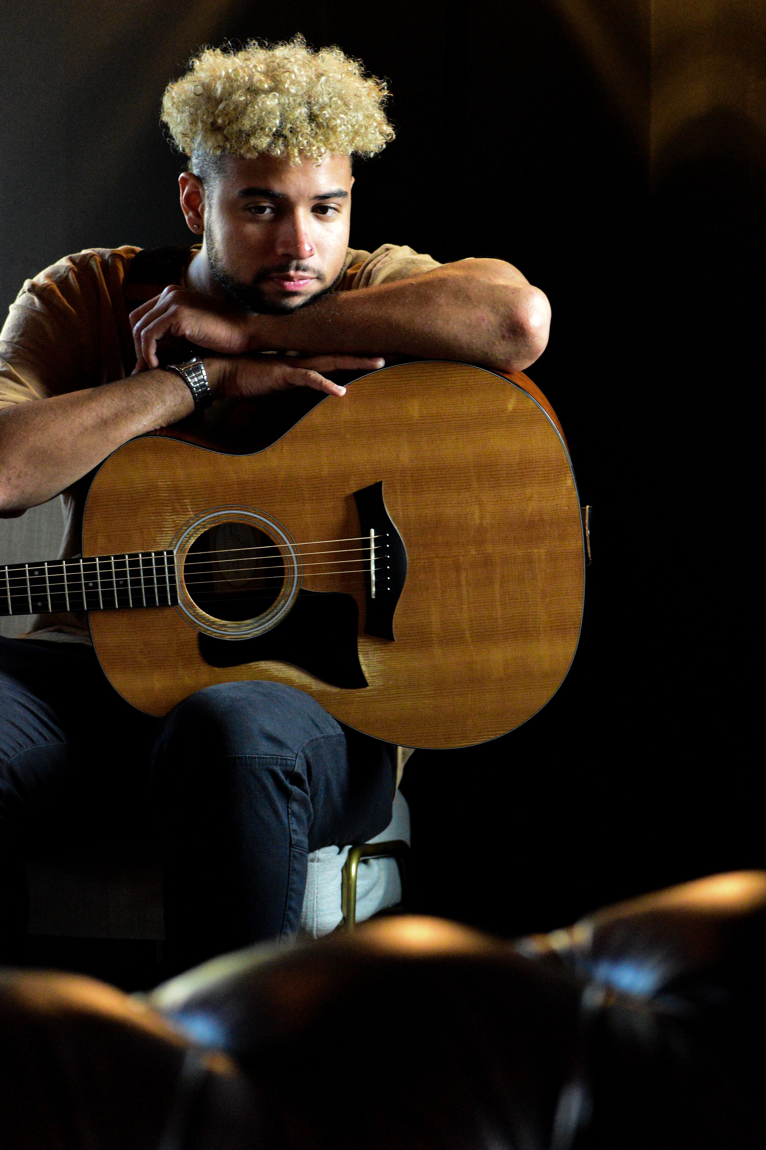 Young man with curly blonde hair resting arms on acoustic guitar in dramatic dark studio setting.