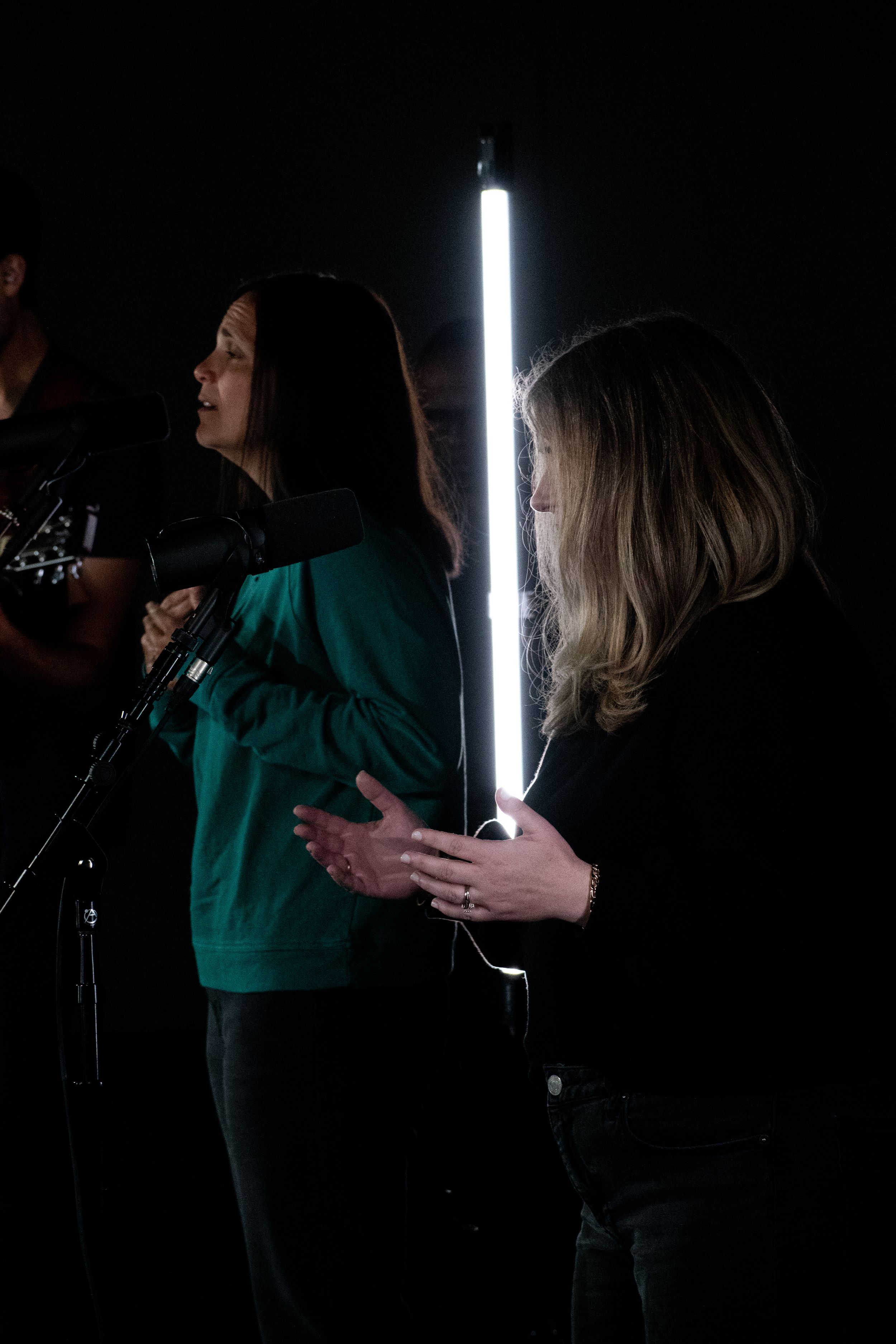 Two women stand in dark studio, one holding glowing LED tube light, wearing green and black tops.
