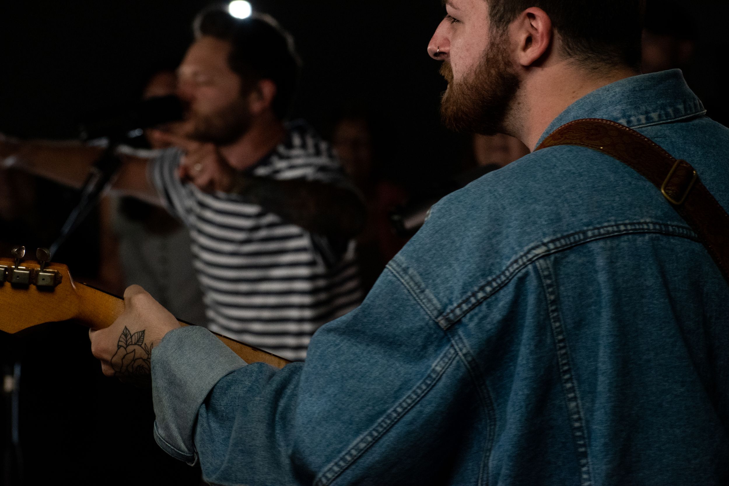 Two bearded musicians performing on stage, one playing guitar in a denim jacket with a tattoo visible.