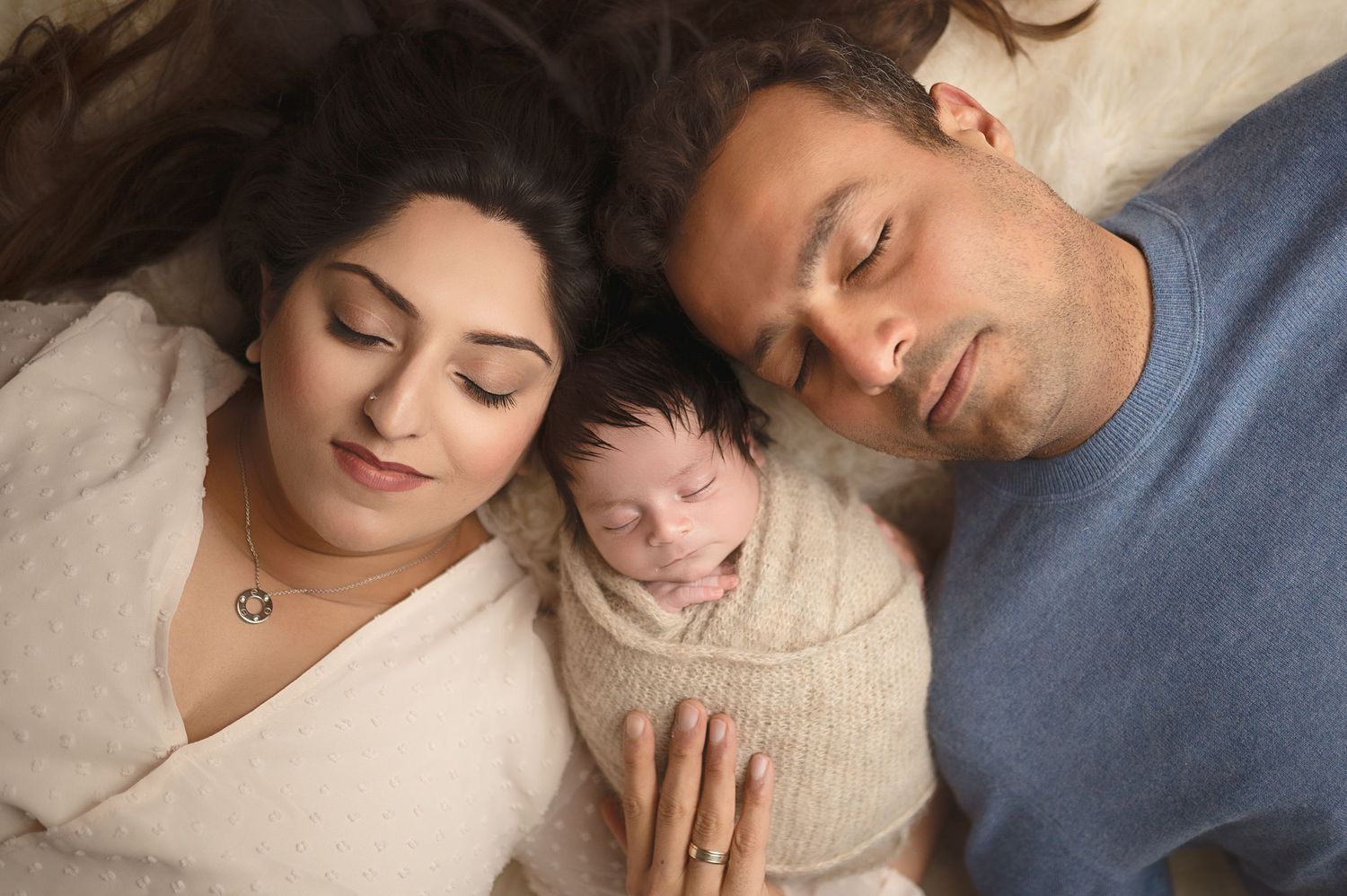 Family portrait with sleeping newborn nestled between parents on light background.