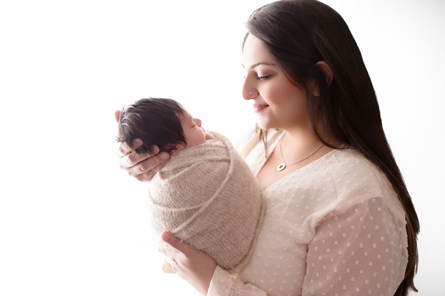 Side profile of parent holding sleeping newborn wrapped in tan swaddle against white backdrop.