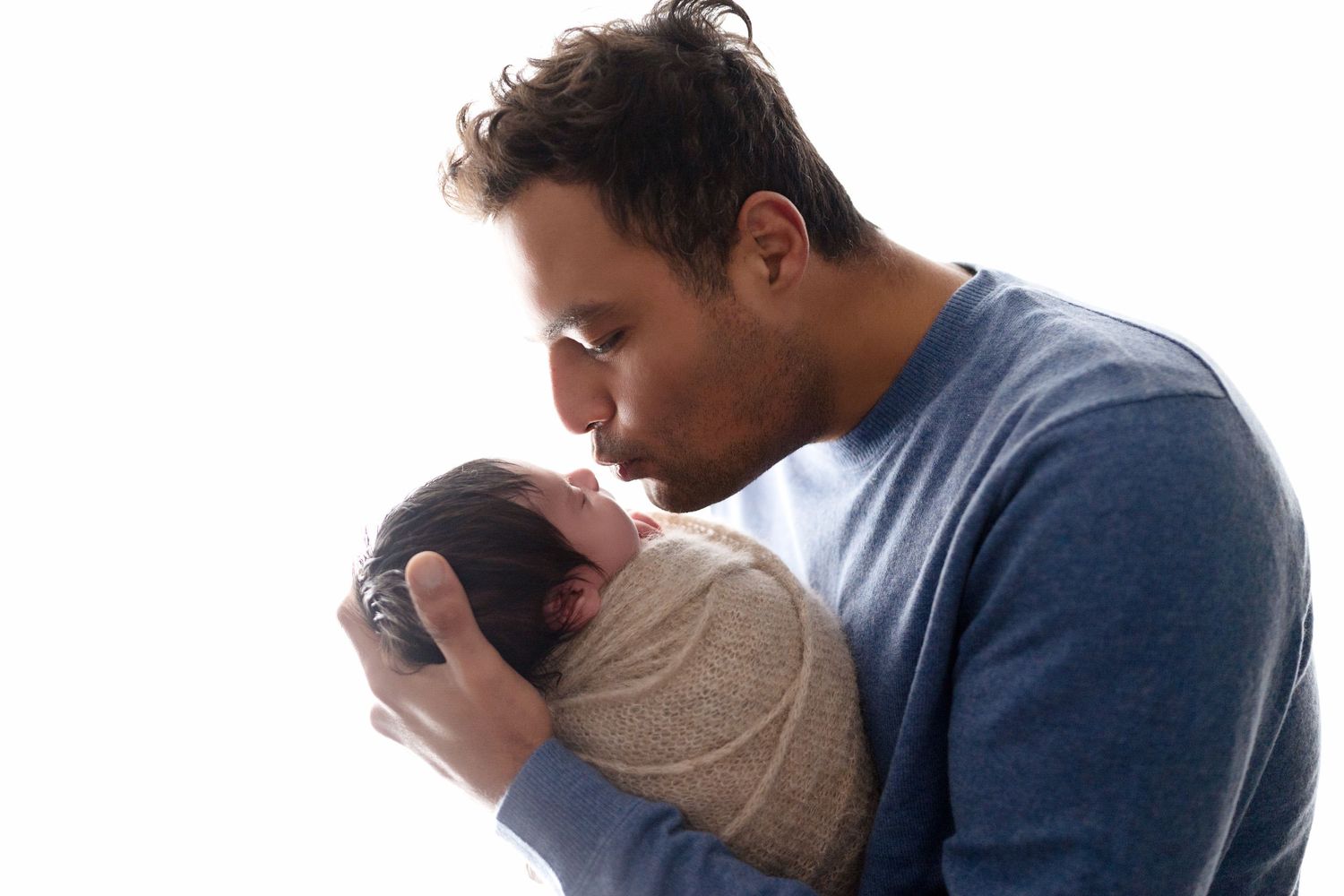 Intimate portrait of parent cradling and kissing newborn wrapped in beige swaddle.