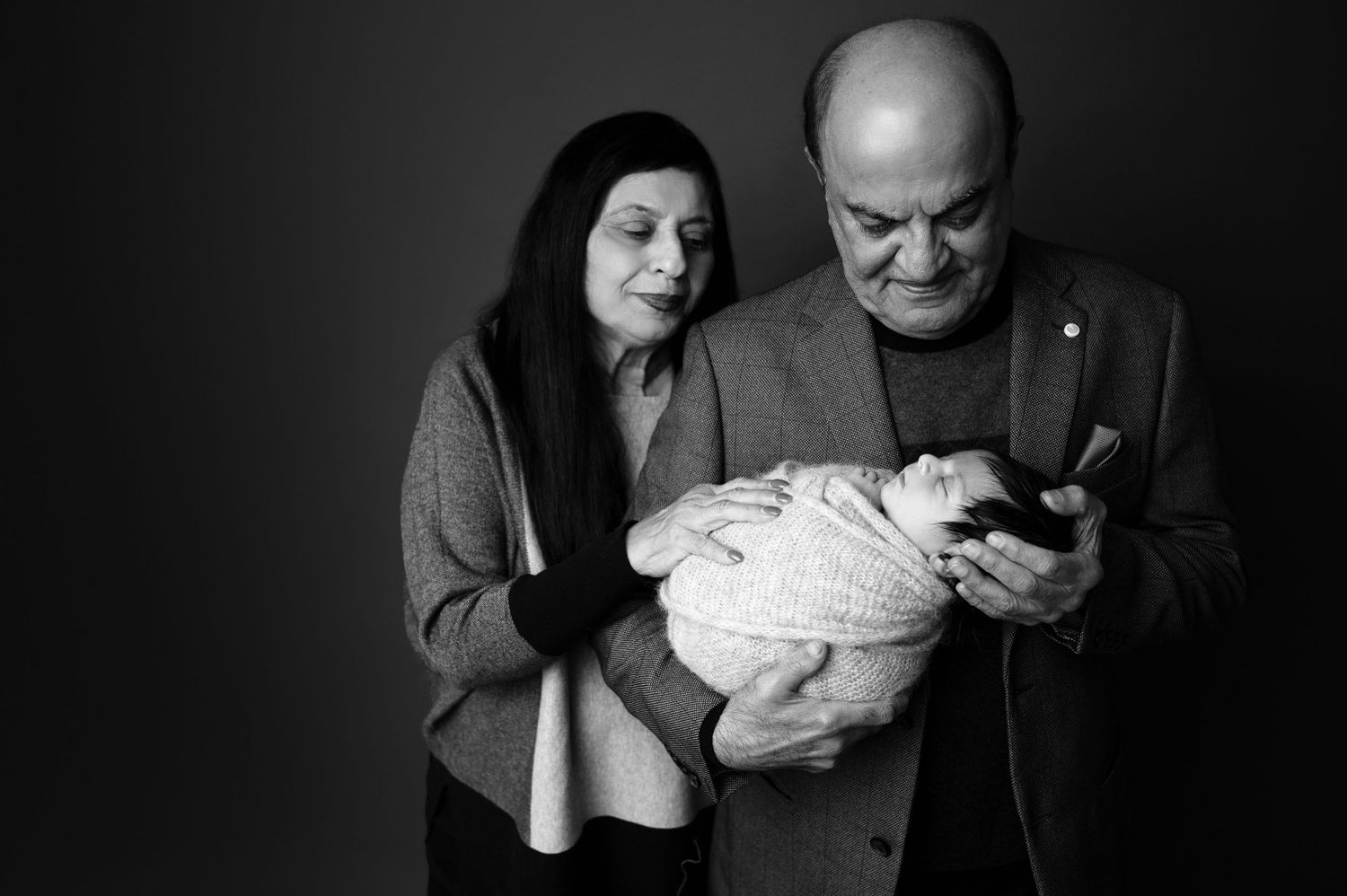 Emotional black and white photograph of adults gazing lovingly at a newborn baby.