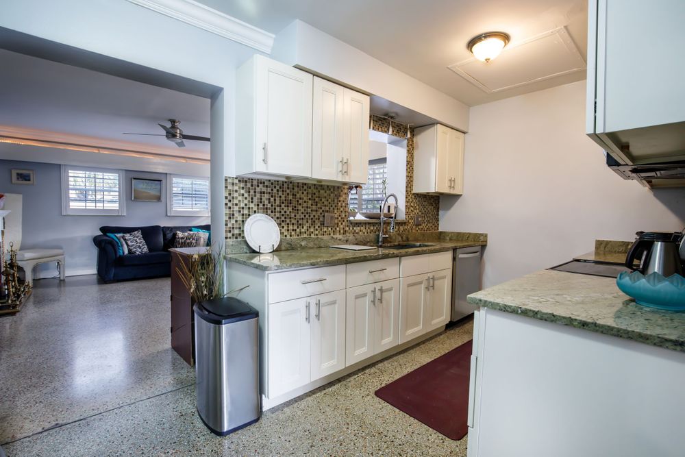 Long view of white kitchen with granite counters and modern appliances leading to living area.