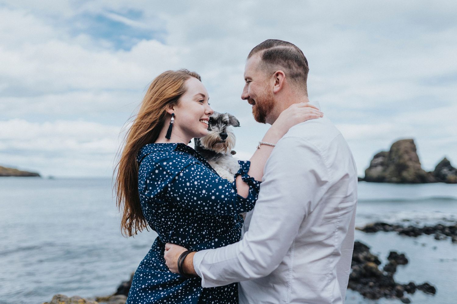 a couple posing with their dog with the sea behind them