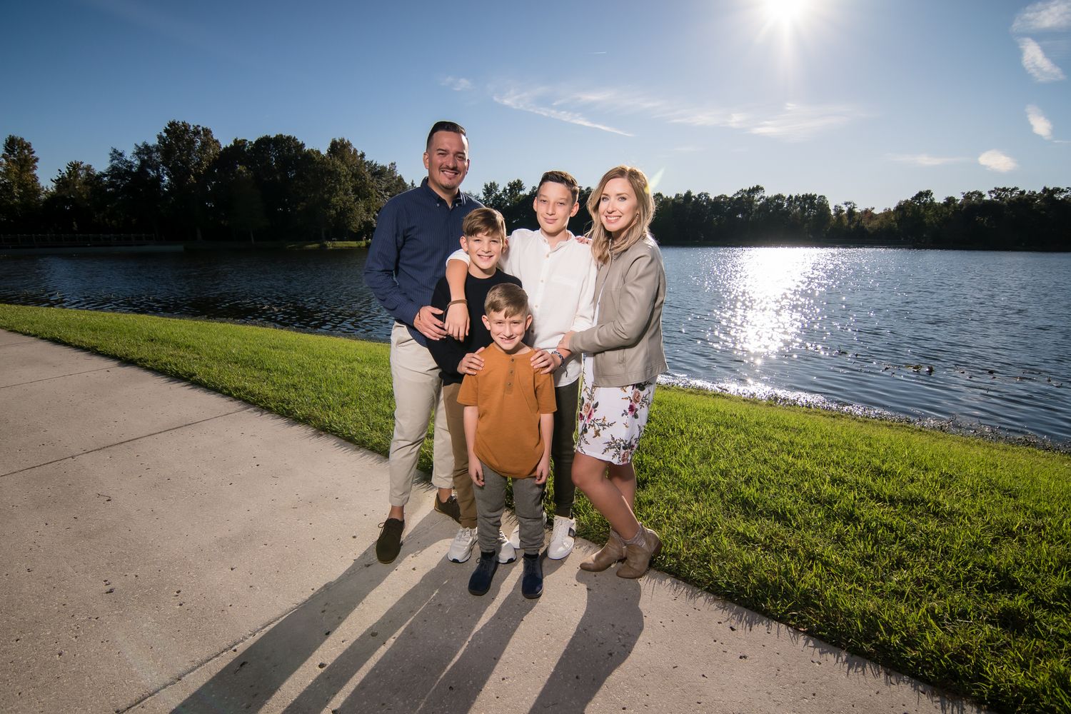 A family stands together by a sunlit lake with reflective water and trees in the background.