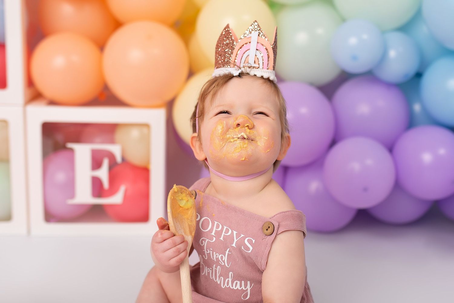 Birthday photo with rainbow balloon wall and sparkly crown against colorful backdrop.
