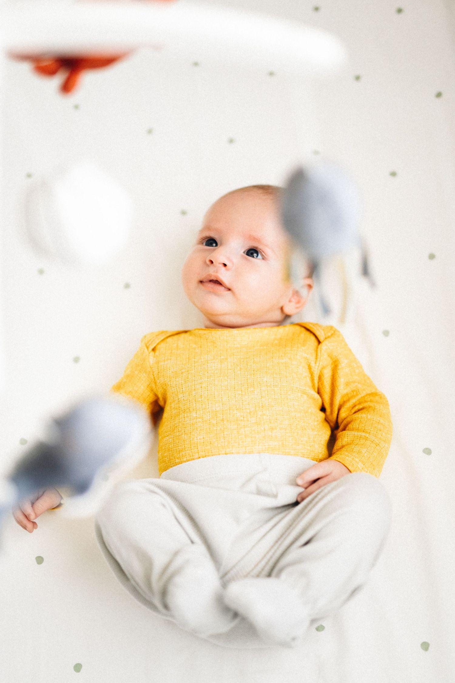 Baby wearing yellow sweater lies beneath mobile toys on dotted background.